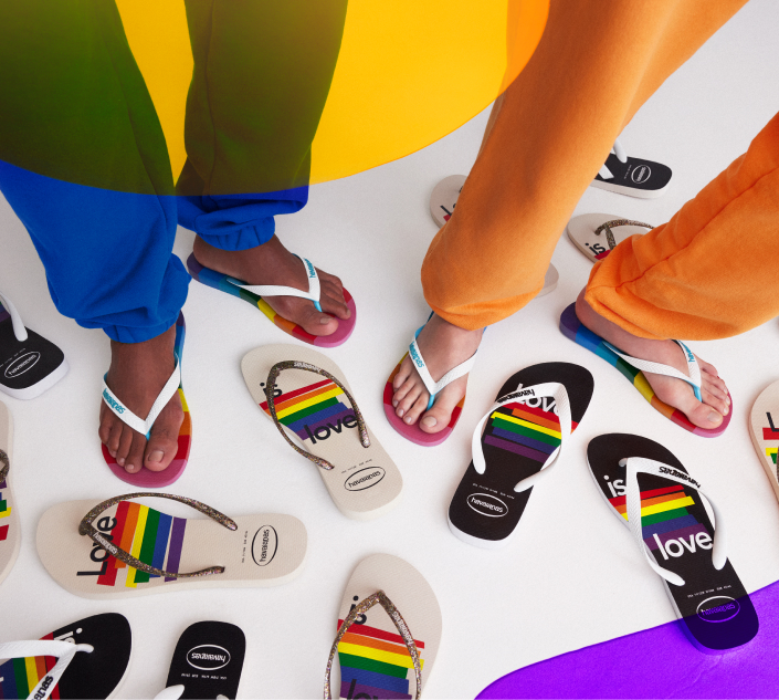 Two people wearing Havaianas pride flip flops. Only the legs and feet are visible. On the white floor there are flip flops scattered around them