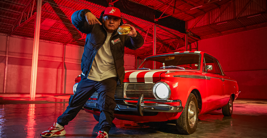 Mexico’s top freestyling artist ‘Aczino’ is standing in front of a red car holding a KFC chicken sandwich