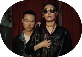 Two people in an urban inside environment. Both are wearing glasses. A guy wearing normal glasses stand aa bit behind a women who is wearing sunglasses with a red cord attachted to it.