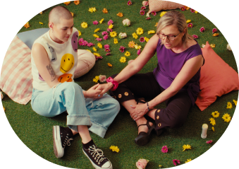 A young women sitting on the ground holding hands with her mother who also sits on the ground. There are cushions and flowers on the ground. It's a colourful image.