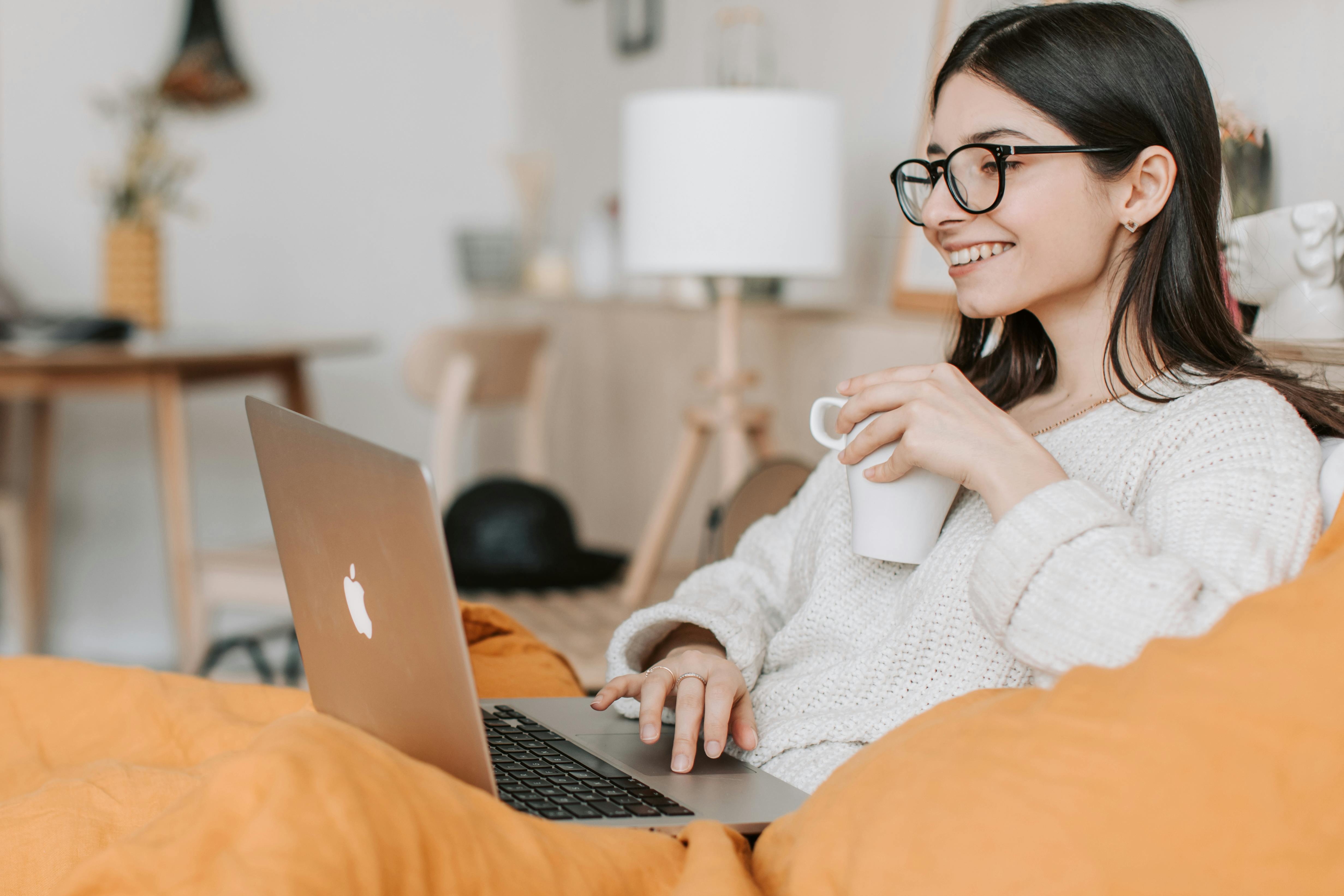 Women sitting on a couch looking at a laptop screen and she has a cup in her hand.
