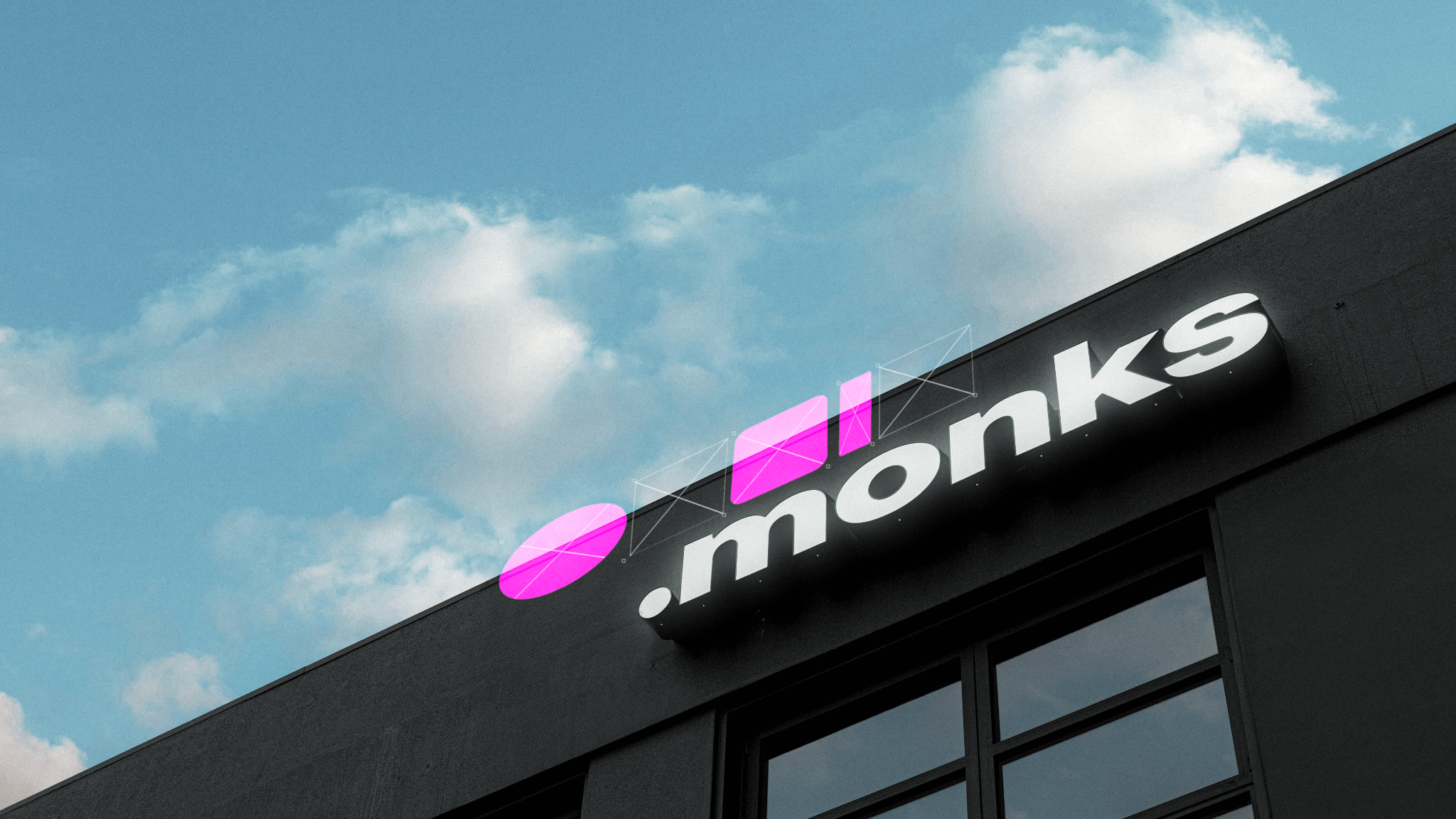 A modern building with a sign that reads ".monks" in white letters on a black background. Above the text are two geometric shapes in pink: a circle and a square. The sky is blue with scattered clouds in the background.