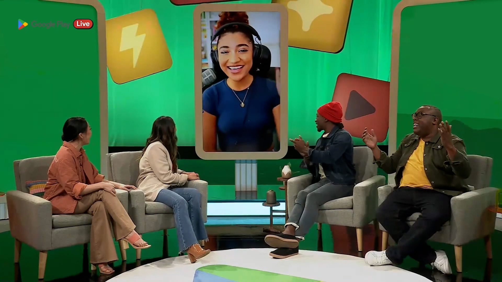 The image shows a lively studio set where four presenters are engaged in conversation, with one presenter appearing via video call on a large screen, all set against a vibrant green backdrop decorated with colorful graphics.