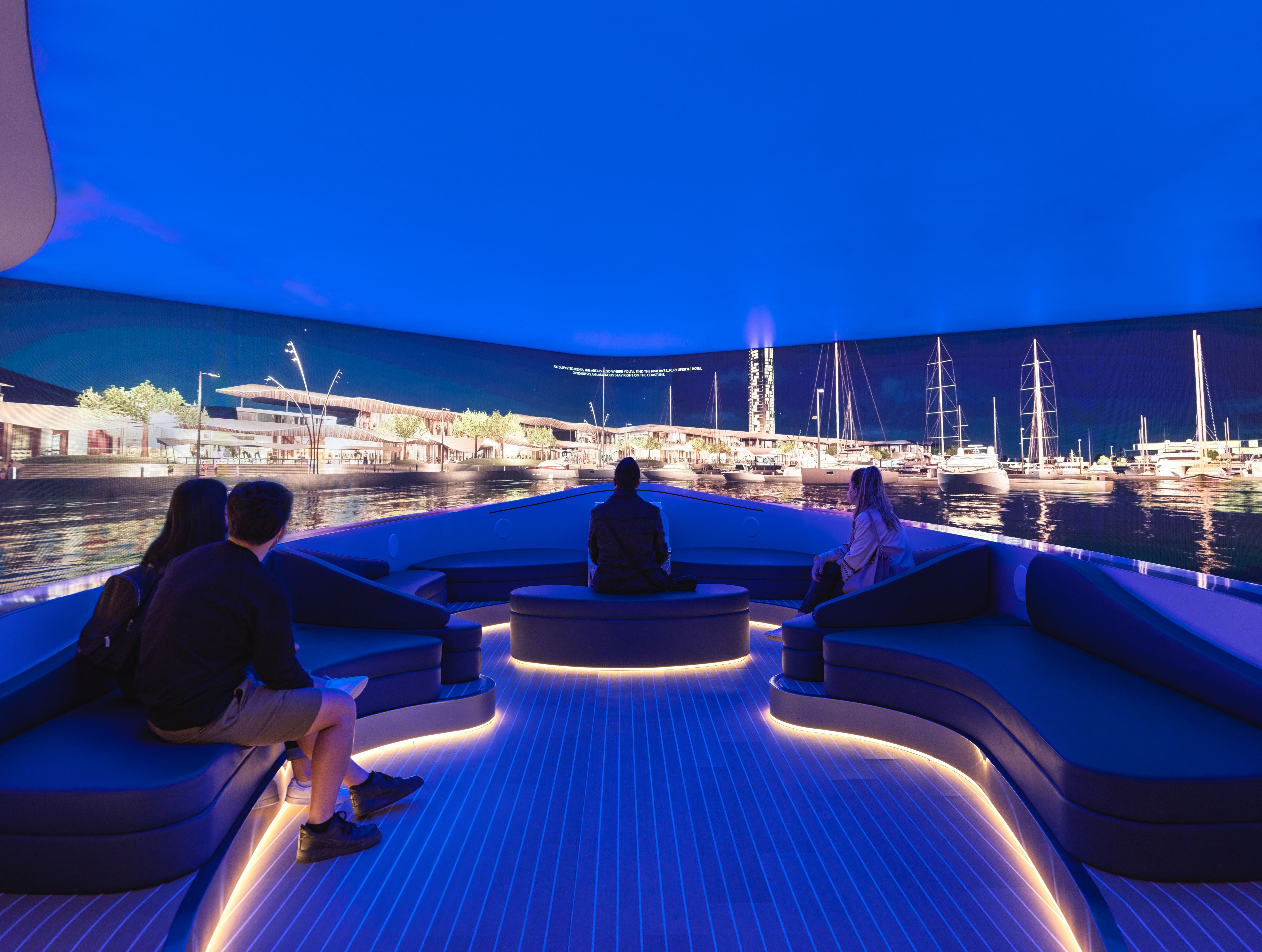 A group of visitors are seated on curved, illuminated benches inside an immersive experience room designed to look like the deck of a yacht. They are watching a panoramic, wrap-around screen displaying a vibrant, photorealistic marina at night.