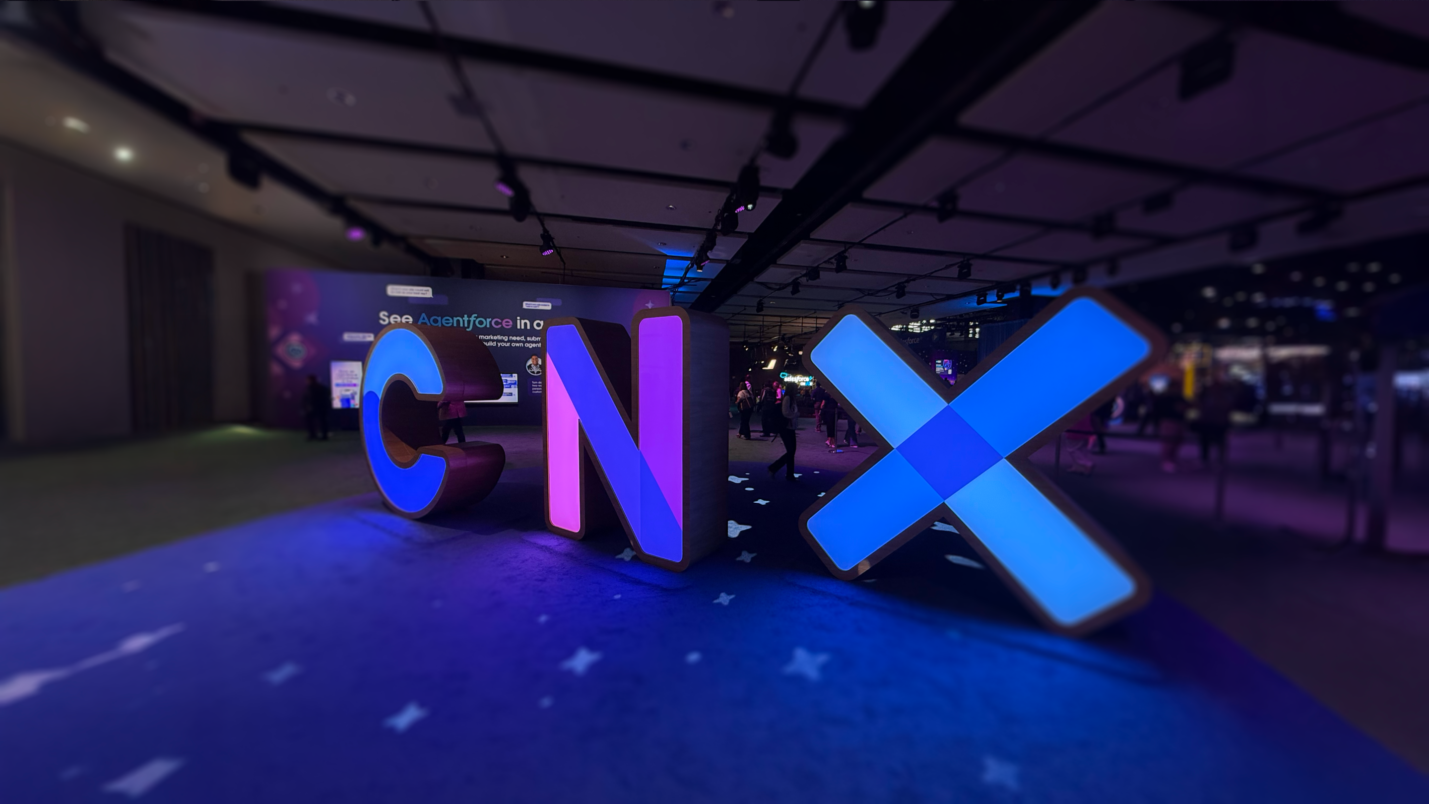 A low-angle, wide shot of large, illuminated, three-dimensional letters spelling "CNX" in a dimly lit event hall. The 'C' glows blue, the 'N' glows purple, and the 'X' glows with a bright blue light. The letters are placed on a dark blue carpet with a subtle pattern, and they cast colorful reflections on the floor. In the blurred background, people can be seen walking around the conference space.