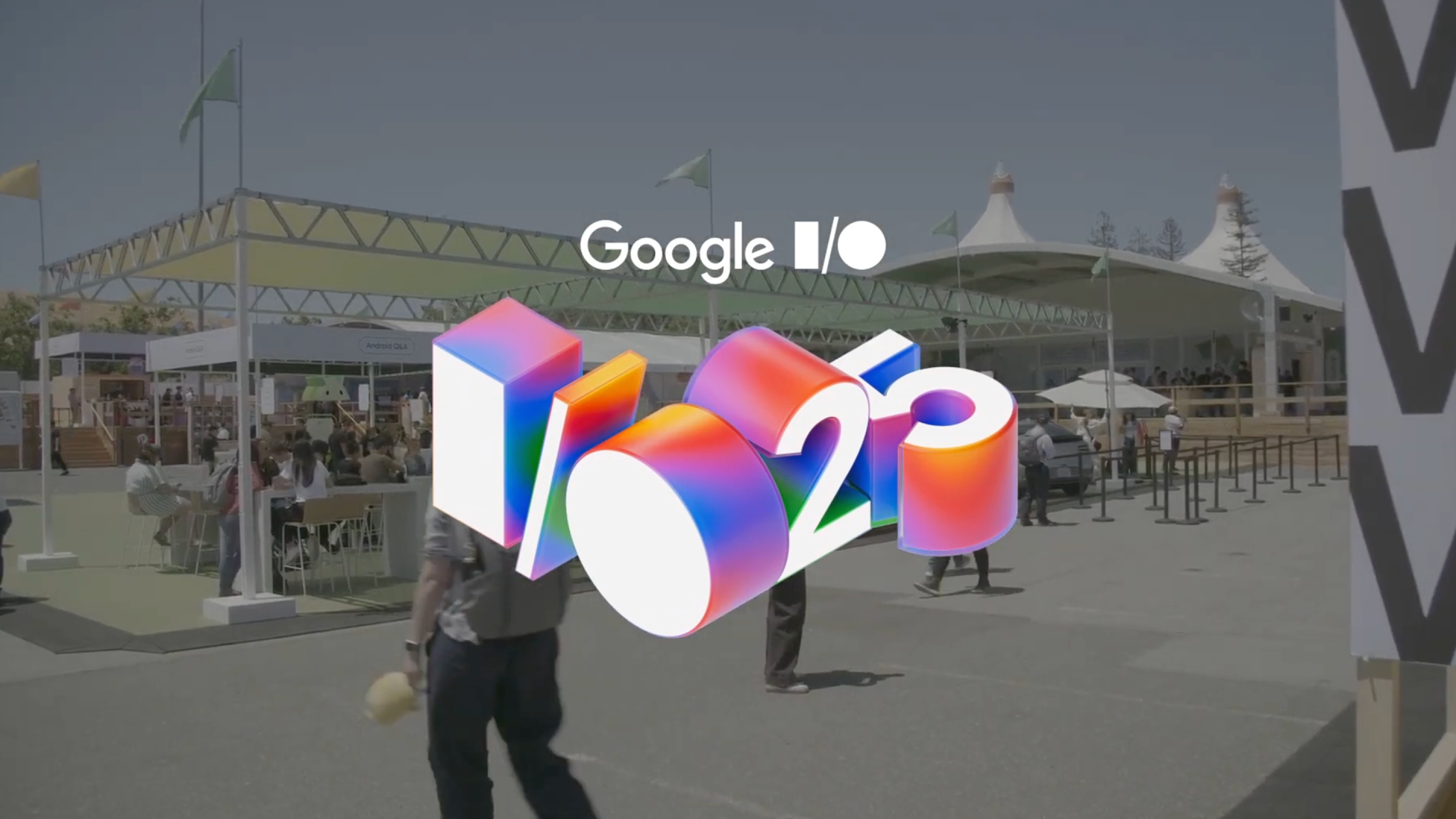 An outdoor shot of the Google I/O 2025 conference on a sunny day. In the foreground is a large, colorful, 3D graphic of the "I/O '25" logo. In the background, attendees walk between large white tents and event structures.