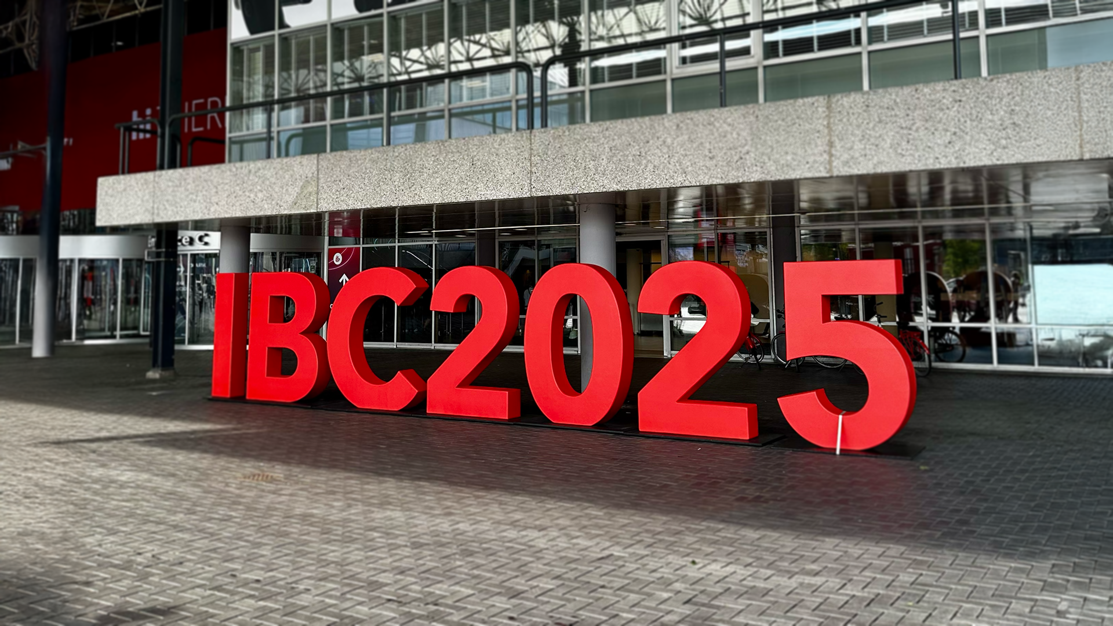 Large, three-dimensional red letters spelling "IBC2025" stand on a brick plaza in front of the entrance to a modern convention center with a glass facade.