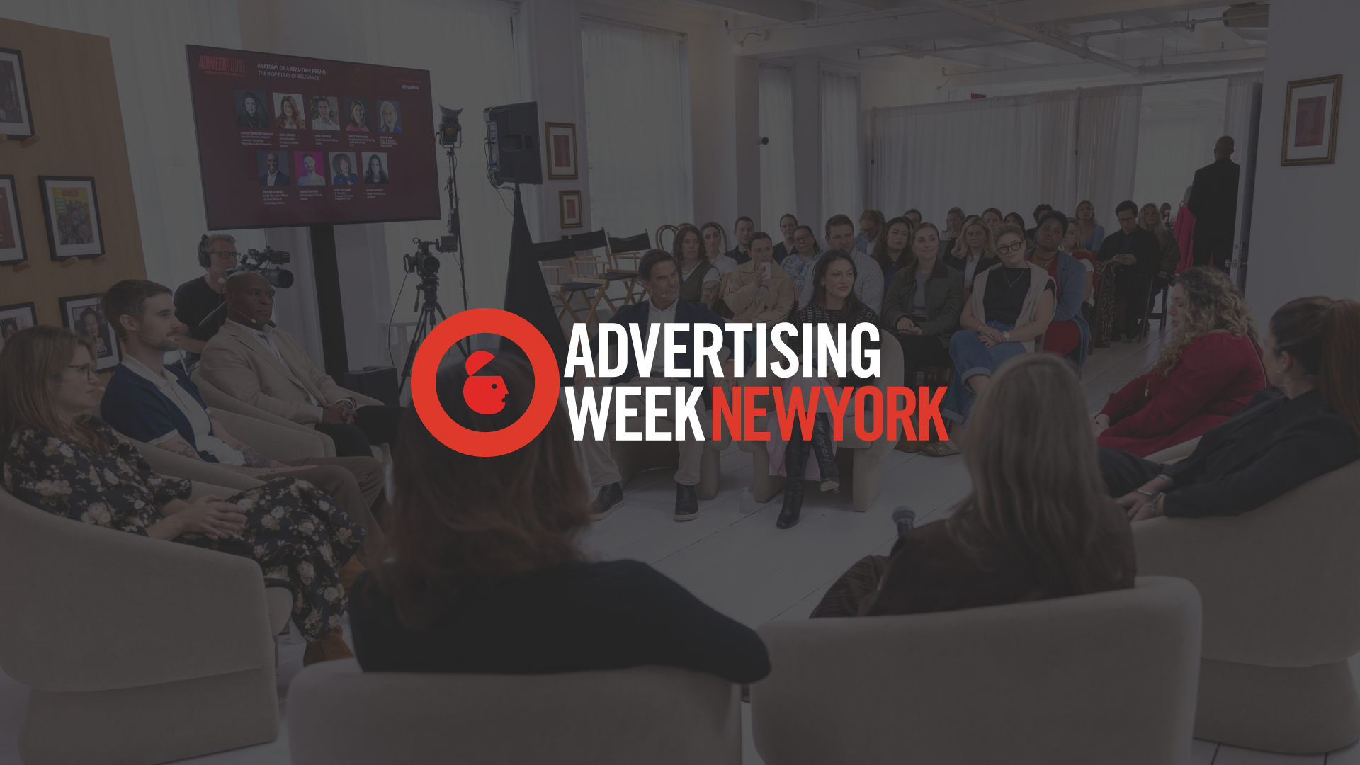 A panel discussion in progress at an event, with the "Advertising Week New York" logo overlaid in the center. In the background, a group of panelists sits in a semi-circle of chairs facing an audience, with a large screen displaying speaker headshots on the wall.
