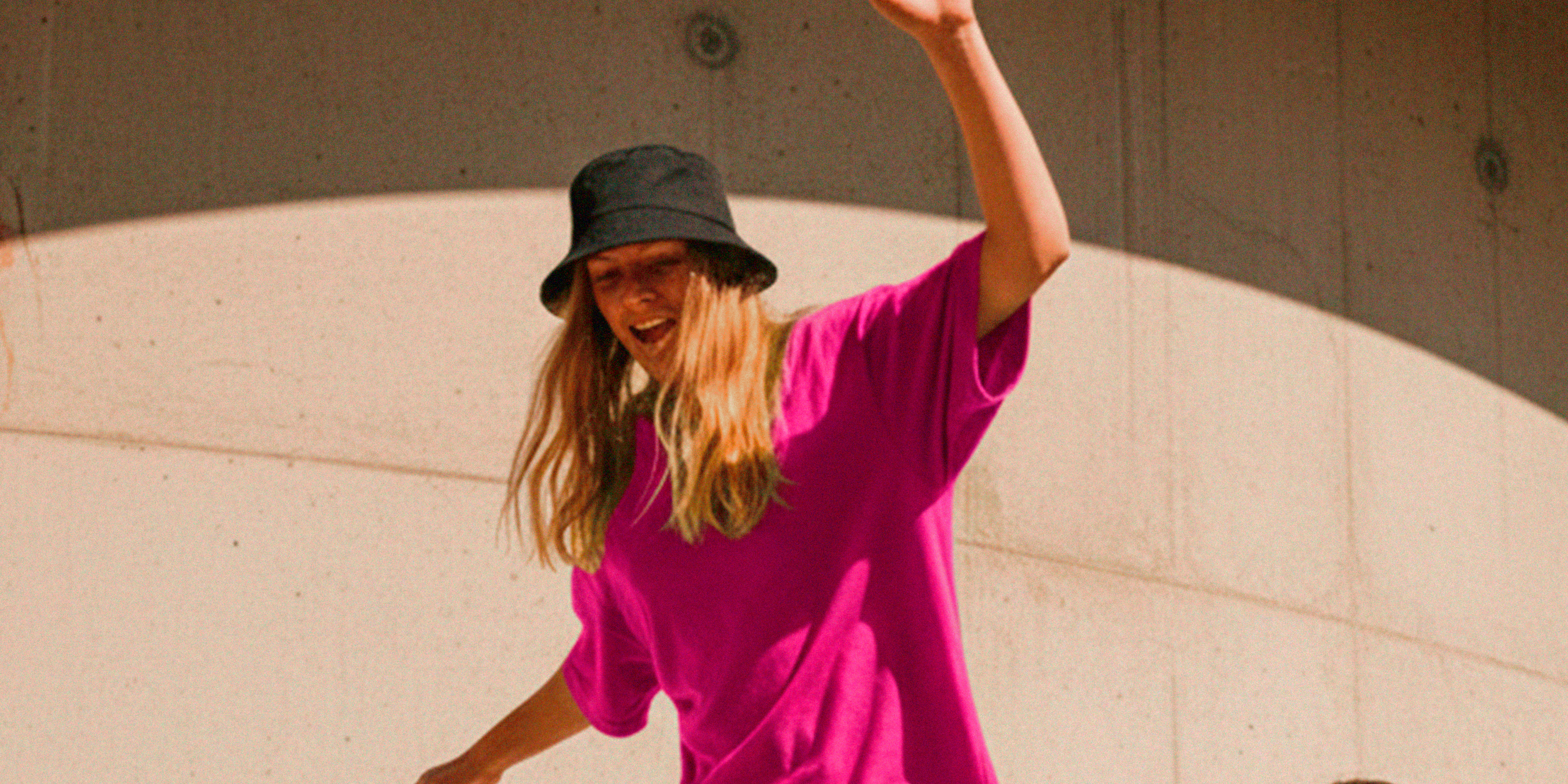 dois jovens em um dia ensolarado. Uma moça de cabelos loiros longos, vestindo uma camiseta oversized rosa-pink, calça clara e chapéu bucket preto, está em pé dentro de um carrinho de supermercado, equilibrando-se com os braços abertos e expressão alegre. Um rapaz ao lado, usando camiseta lilás, calça clara e tênis brancos, empurra o carrinho sorrindo. A cena acontece em um ambiente urbano, com fundo de parede de concreto curvada e uma grande sombra semicircular projetada pela luz do sol. 
