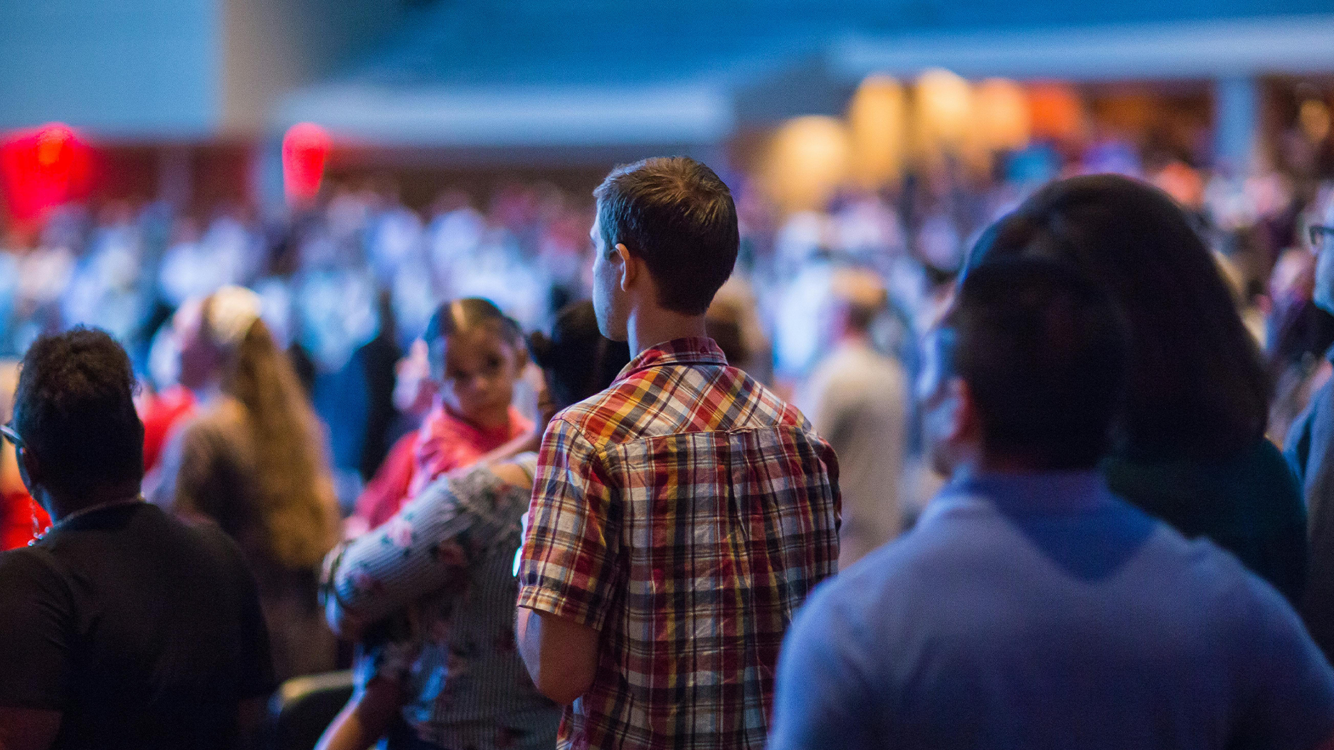 A rear view of a standing crowd in a large, dimly lit indoor venue, focusing on a man in a plaid shirt in the center. To his left, a woman holds a small child who is looking sideways, while the foreground and background contain other attendees blurred out of focus.
