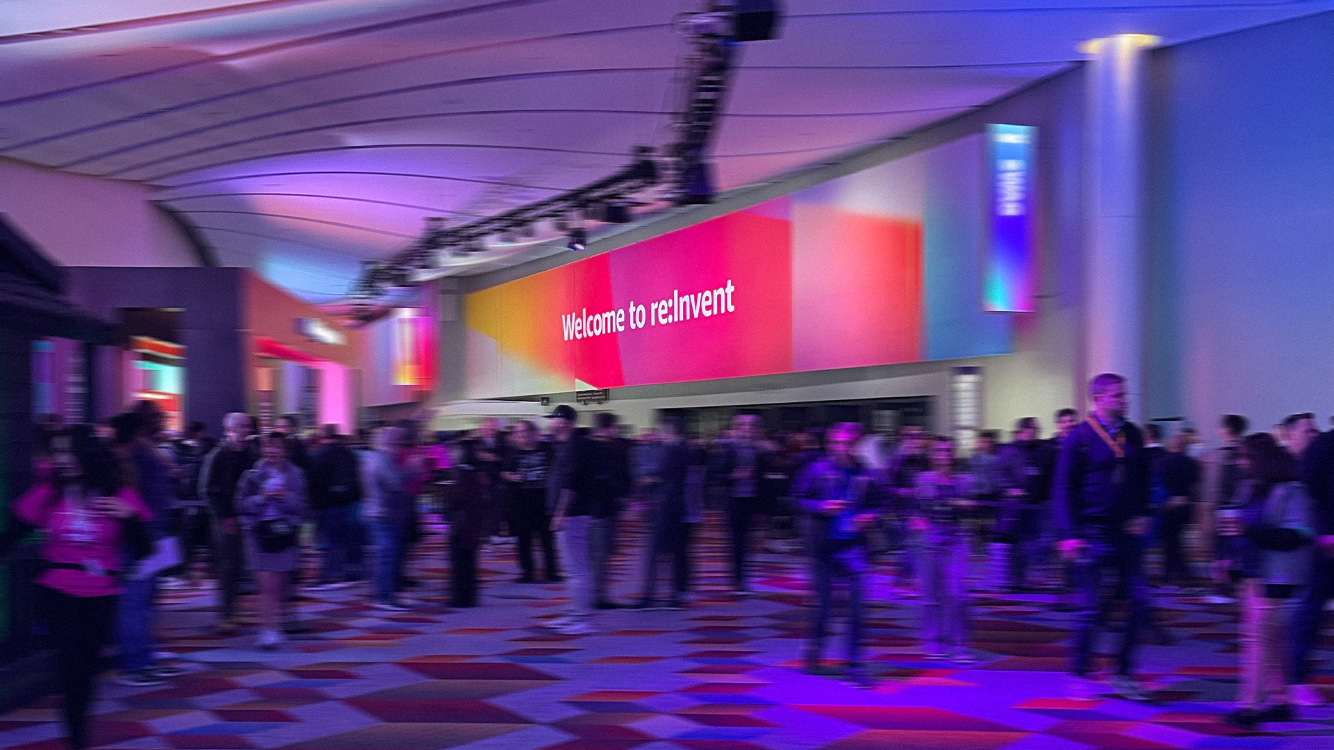 A photograph of a large, crowded convention center hall. A large, curved sign with a colorful pink, orange, and purple gradient background reads "Welcome to re:Invent". The space is illuminated with purple and blue lights, and the floor has a geometric pattern. Numerous attendees are walking around the hall.