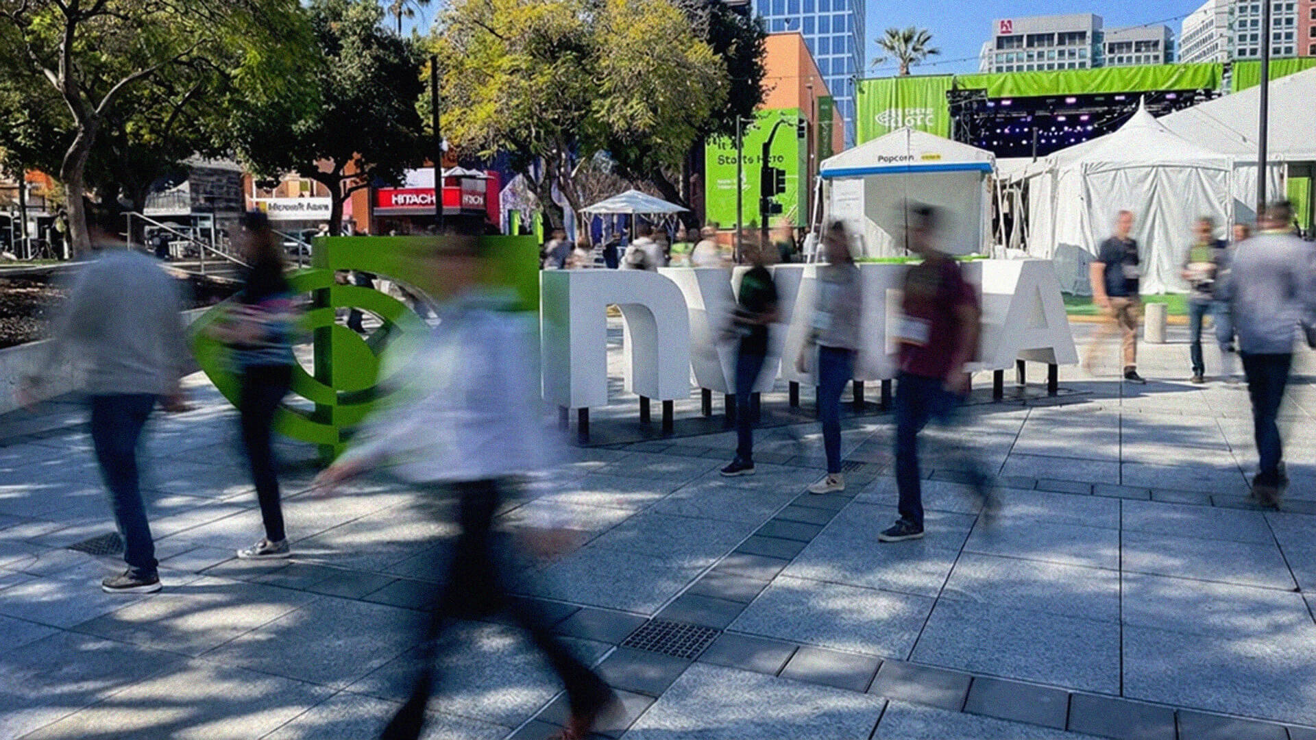 A wide-angle, slightly blurred shot of an outdoor plaza at the NVIDIA GTC 2026 conference in San Jose. Large, 3D white letters spelling out "NVIDIA" stand in the center, with the green NVIDIA logo to the left. People are captured in motion, appearing as blurred figures walking across the stone-tiled ground, creating a sense of a busy, electric atmosphere. In the background, there are green banners, white event tents, trees, and city buildings under a clear blue sky.