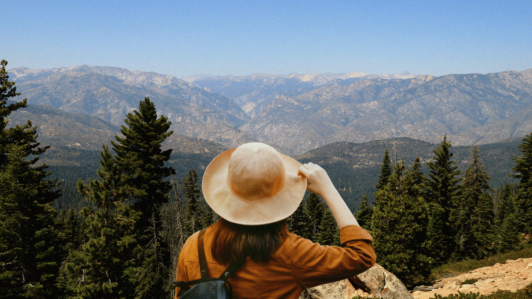 A person seen from behind wearing a wide-brimmed tan hat and an ochre jacket, gazing out at a vast, rugged mountain range and evergreen forest under a clear blue sky.
