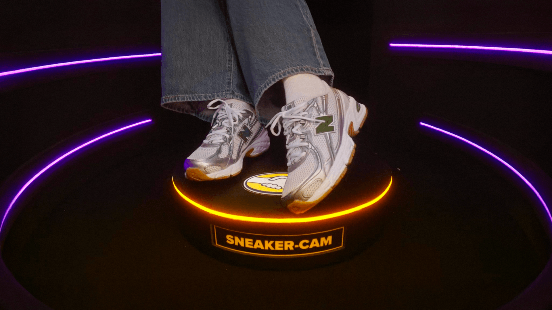 A close-up of a person's feet wearing blue jeans, white socks, and silver and white New Balance sneakers featuring a green "N" logo. They are standing on a small, circular black platform that has a glowing orange ring light around its top edge. The front of the platform displays a yellow label reading "SNEAKER-CAM".