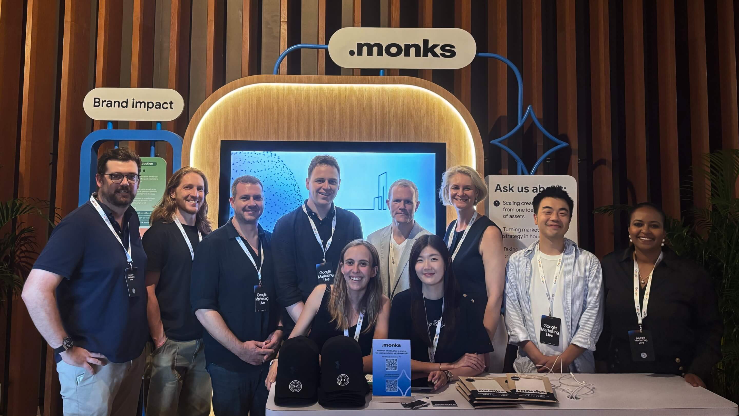 A group of ten smiling professionals posing together at a ".monks" event booth. The attendees are wearing lanyards with "Google Marketing Live" badges. Behind them is a large display screen, an illuminated ".monks" logo, and booth signs reading "Brand impact" and "Ask us about:". In the foreground, a table is set up with branded black caps, brochures, and a small sign featuring a QR code.
