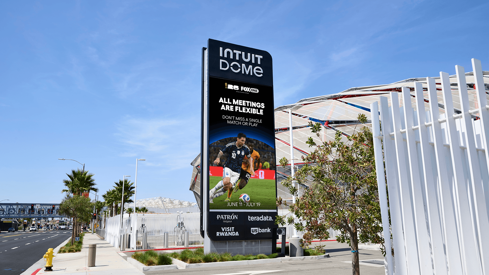 A tall digital billboard outside the Intuit Dome displaying a FIFA World Cup advertisement featuring soccer player Lionel Messi in action. The screen reads "ALL MEETINGS ARE FLEXIBLE" above the dates June 11 through July 19, alongside logos for Fox and various corporate sponsors like Patron and US Bank. The sign stands on a sunny, palm-tree-lined sidewalk with the stadium's exterior visible in the background behind a white fence.