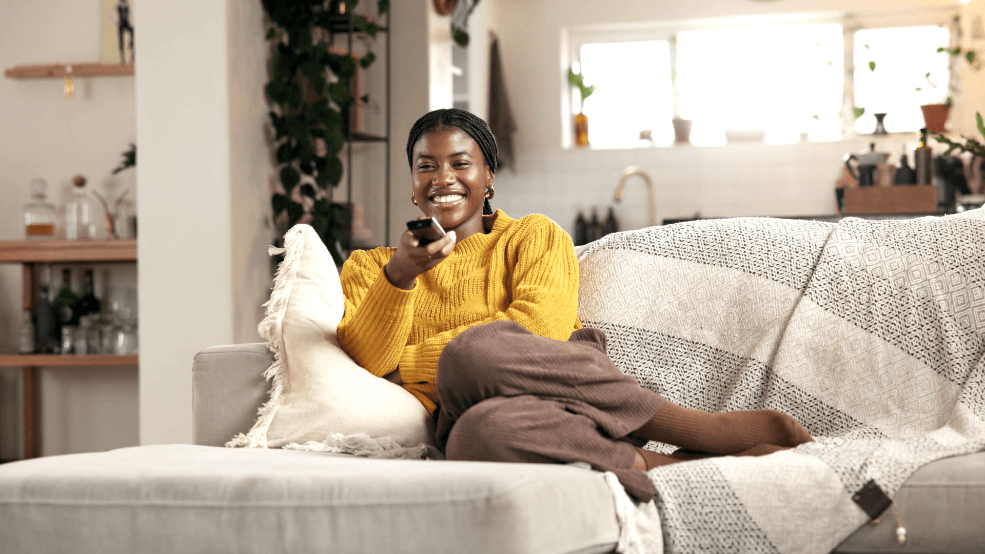 A smiling woman in a yellow sweater relaxes on a couch, holding a TV remote and looking toward the camera.