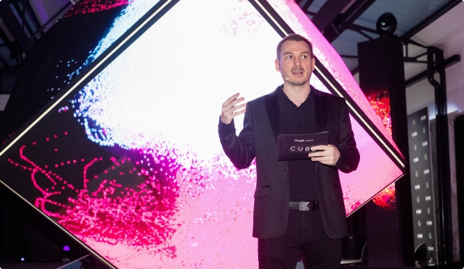 In the image, a man in a black suit is standing in front of a large projection screen. The screen is prominently displayed against a pink background. The man is holding a cell phone, specifically a Samsung Galaxy S10e with a pixelated screen. He is also holding a microphone, indicating that he may be speaking or presenting at an event. The man in the black suit is wearing a mustache and is surrounded by various objects and individuals. There is a small text in the image that reads "CUB" on the lower portio