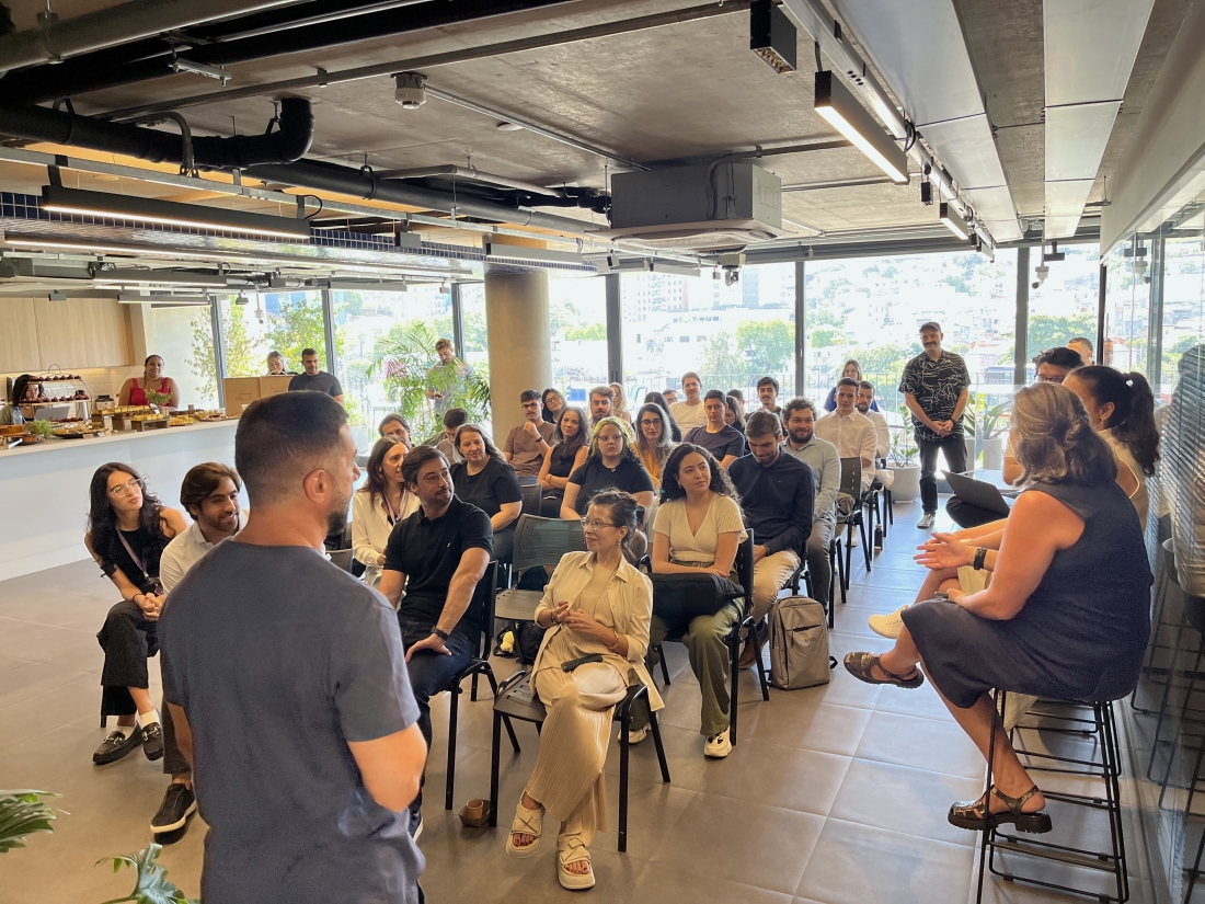 Um ambiente corporativo onde uma pessoa está falando na frente de um grupo diversificado de ouvintes sentados e em pé. A sala é espaçosa e bem iluminada, com grandes janelas ao fundo que oferecem vista para alguns edifícios. A interação parece ser parte de uma reunião informal ou de uma sessão de brainstorming, com um clima confortável e participativo.