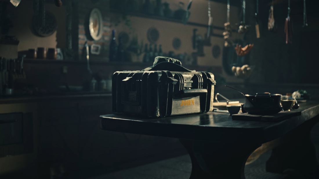 A dark setting of a kitchen table. on that tabel is a dark chest with the name 'John' written on it.