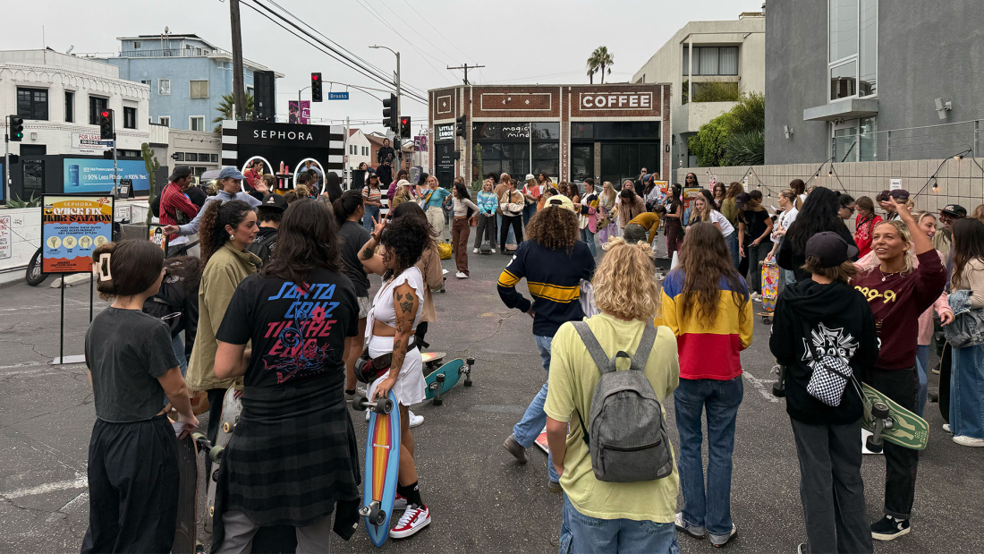 The image shows a lively urban scene bustling with a crowd gathered near a Sephora booth, where various people, including skateboarders, mingle and engage in activities. Bright signage and a nearby coffee shop contribute to the vibrant atmosphere of this community event.