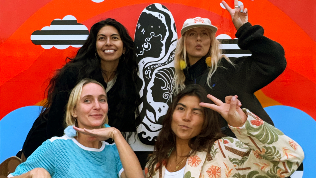 The image shows two smiling women posing in front of a vibrant mural, with one displaying a skateboard and both making playful hand gestures.