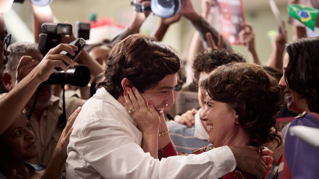 The image depicts two women joyfully embracing amidst a crowd, surrounded by photographers and supporters, conveying a sense of celebration and connection.