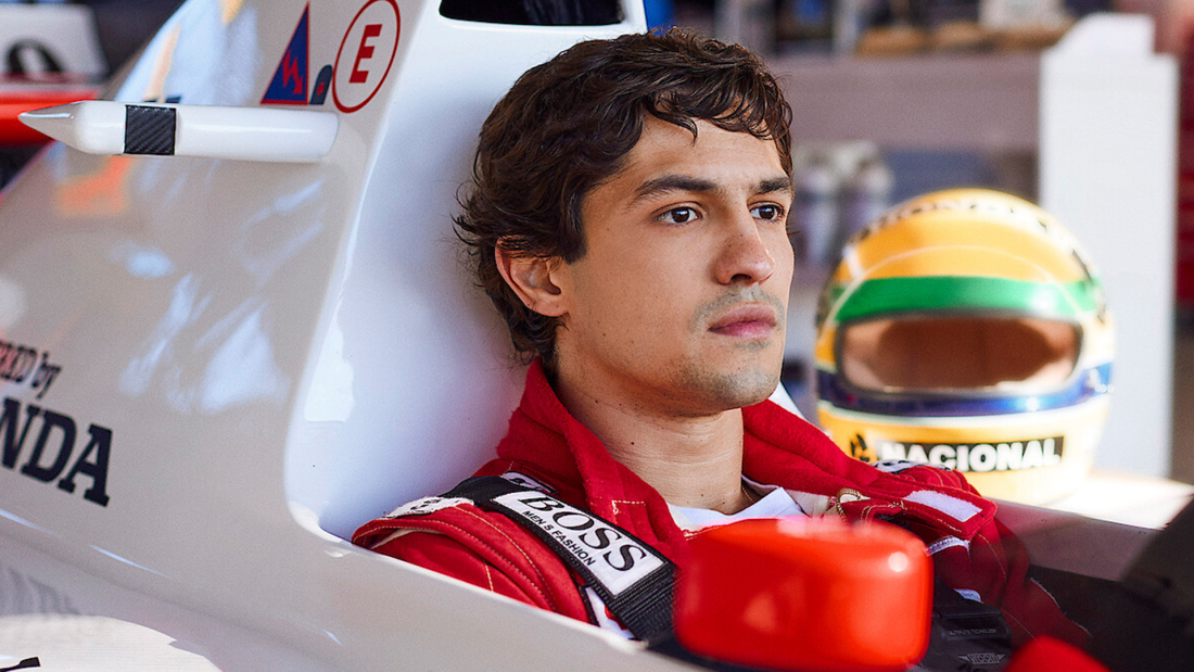 The image features a focused young race car driver seated in a racing cockpit, wearing a red jumpsuit, with a helmet displaying a vibrant design visible in the background.