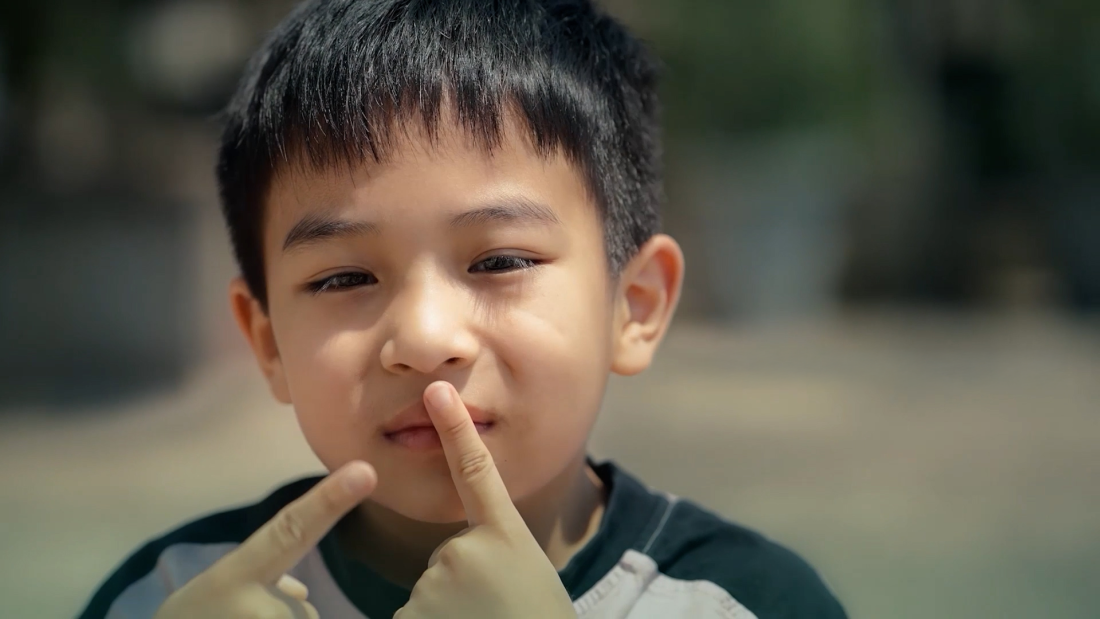 The image shows a young boy outdoors with a thoughtful expression, touching his lips with his finger.