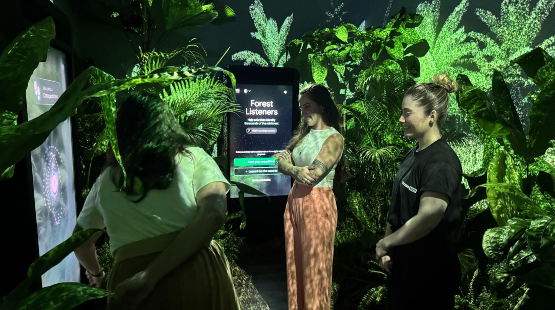 Three women interact with an immersive digital exhibit in a dimly lit room filled with lush tropical plants. A central vertical screen displays the title "Forest Listeners" and invites visitors to help scientists identify rainforest sounds.