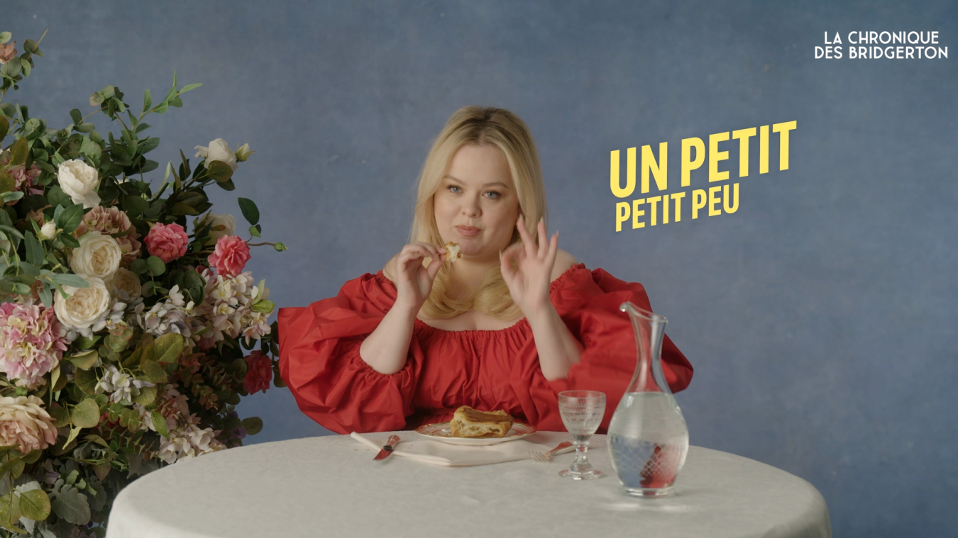 A woman in a bright red dress sits at a table adorned with flowers, holding a piece of food and gesturing, with the text "Un petit petit peu" beside her.