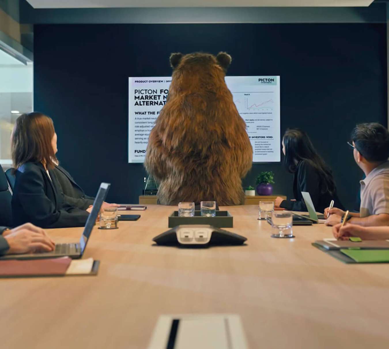 A large bear in an office meeting standing at the front of the meeting table while the other workers are viewing the slide on the TV.