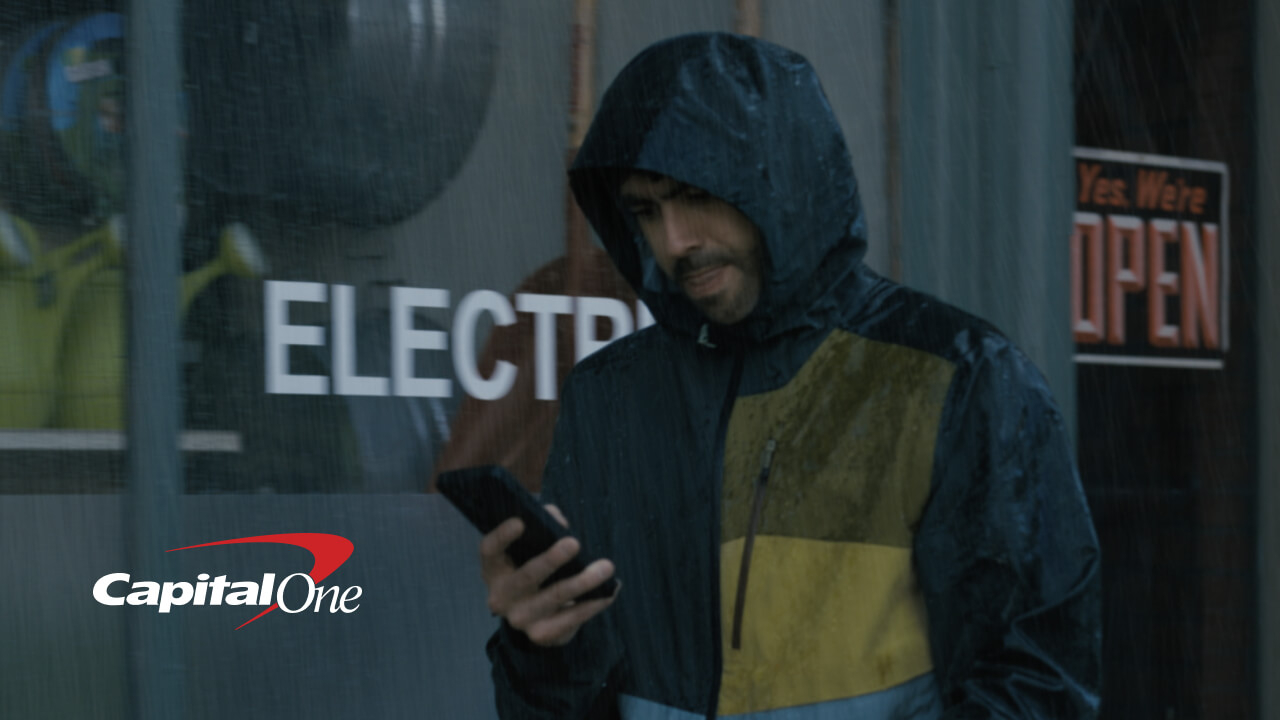 A man wearing a hooded, color-blocked (dark blue and yellow) rain jacket stands outdoors in the rain, looking down at his smartphone. He is standing in front of a storefront window that has the partial word 'ELECTR' on the glass and a 'Yes, We're OPEN' sign hanging nearby. The Capital One logo is displayed in the bottom left corner.