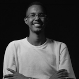 Foto em preto e branco de um homem sorridente, usando óculos e uma camiseta clara. Ele tem cabelo curto e barba curta, e está posando com os braços cruzados. O fundo da imagem é escuro, destacando o rosto e a expressão acolhedora do homem.