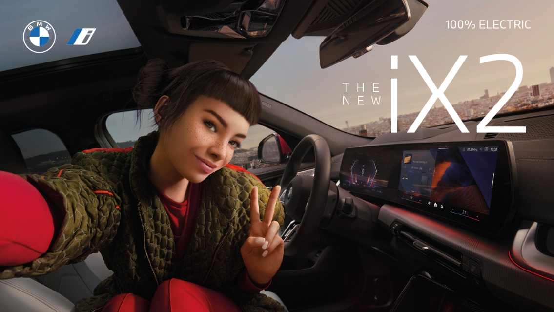 You see the interior of the BMW iX2 car. Miquela dressed in a red pants is sitting on the driver car and makes a peace sign with her hand.
