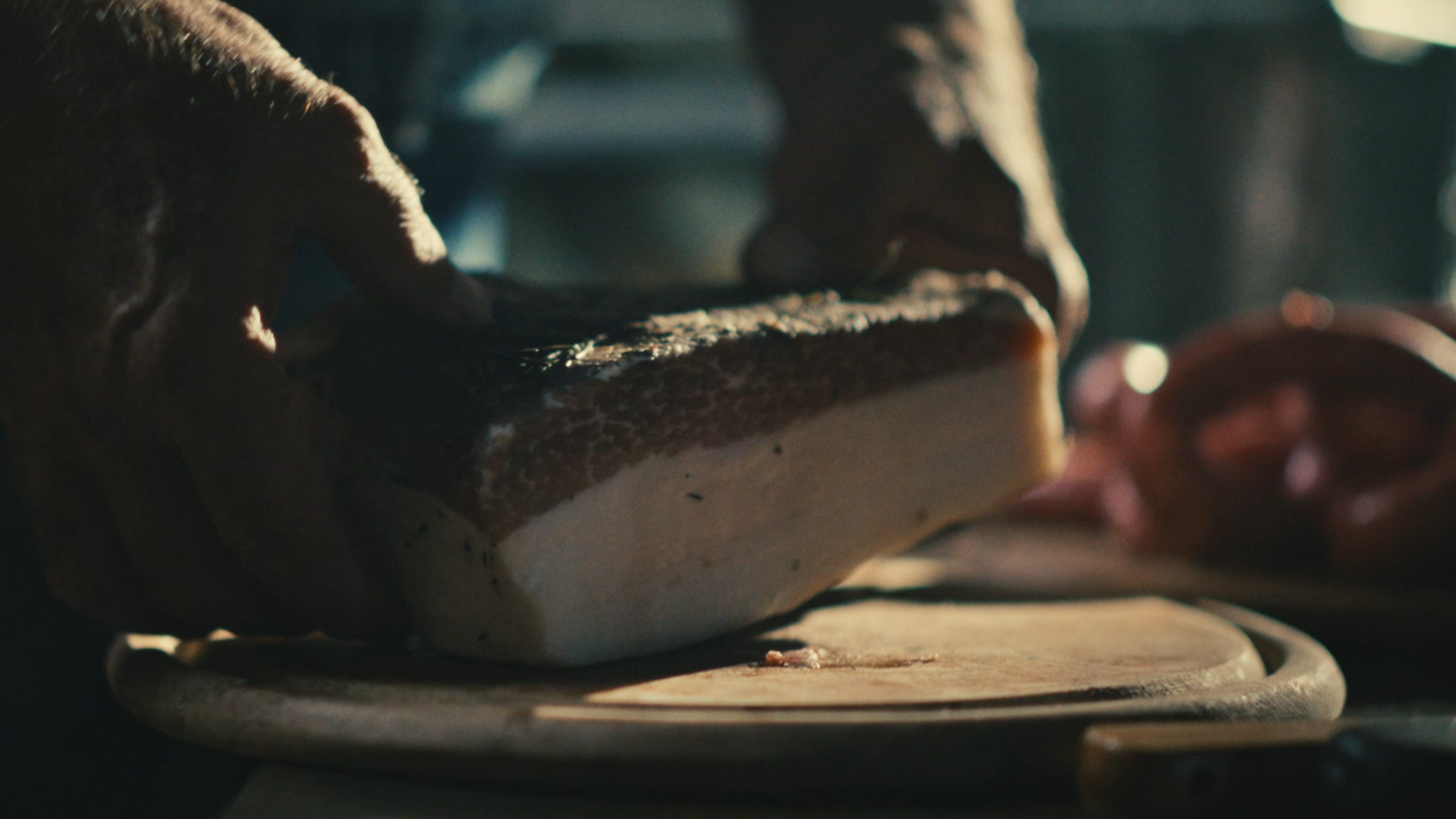 Hands that are putting a big ham on a wooden plank on a kitchen table.