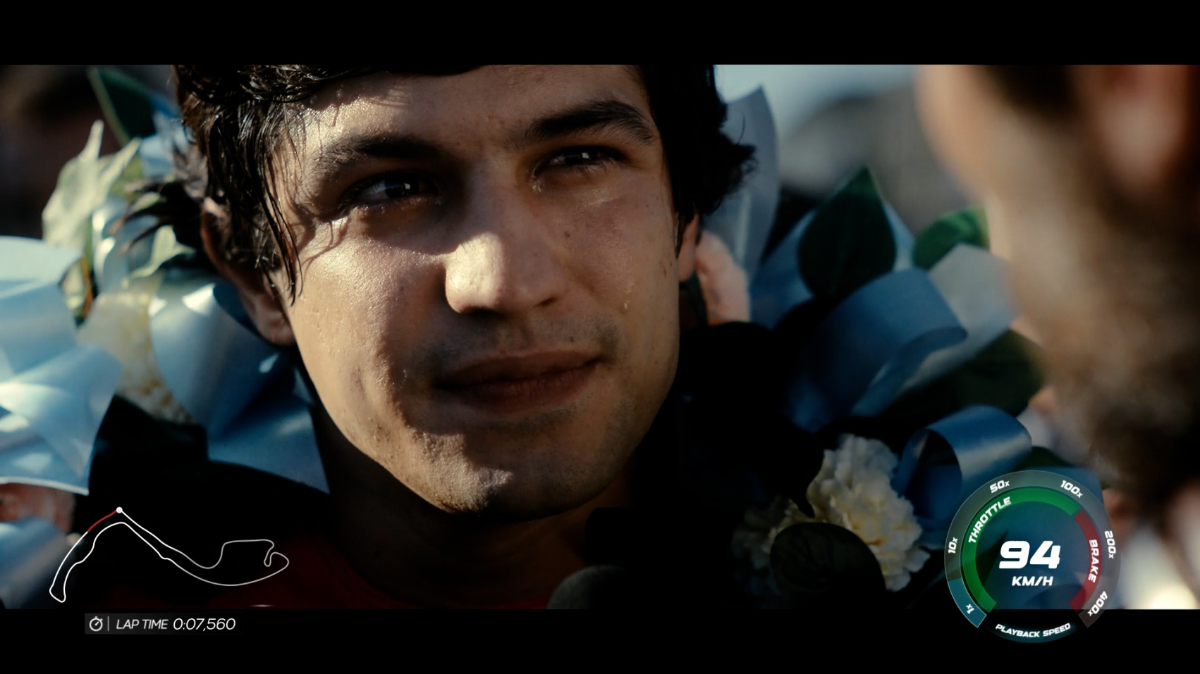 The image captures a close-up of a race car driver adorned with a floral wreath, visibly emotional with a tear on his cheek, as he interacts with another individual, set against a racing backdrop.