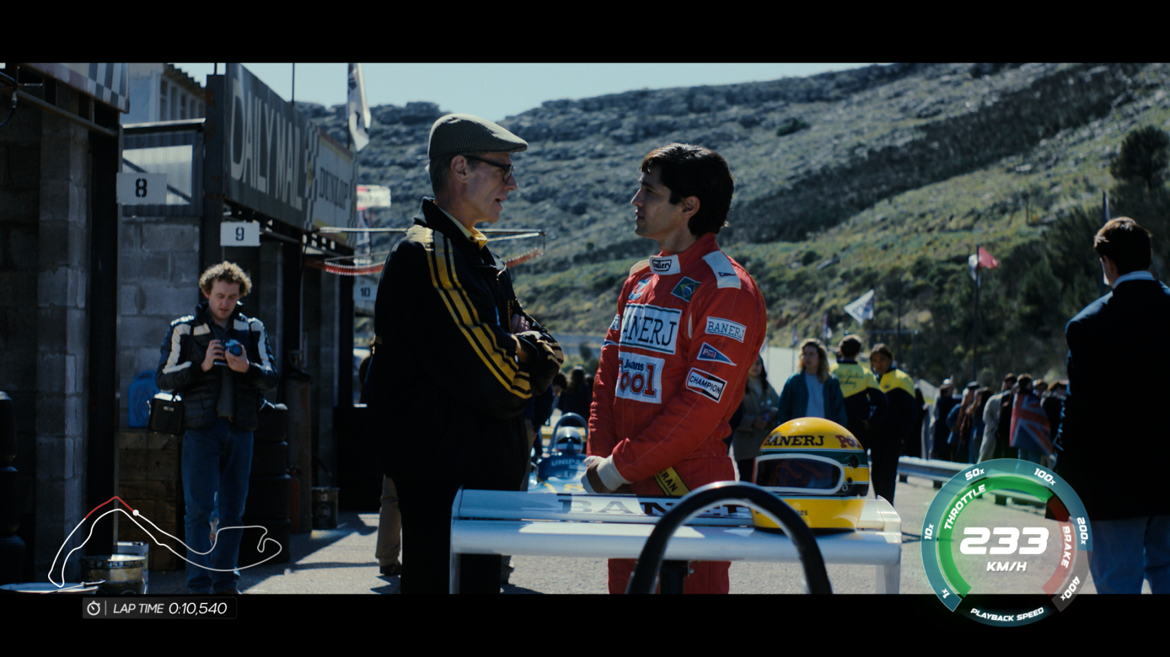 The image shows a serious interaction between a race car driver in a red suit and an older man in a cap, set against a backdrop of racing pits and spectators, suggesting a significant conversation in a racing environment.