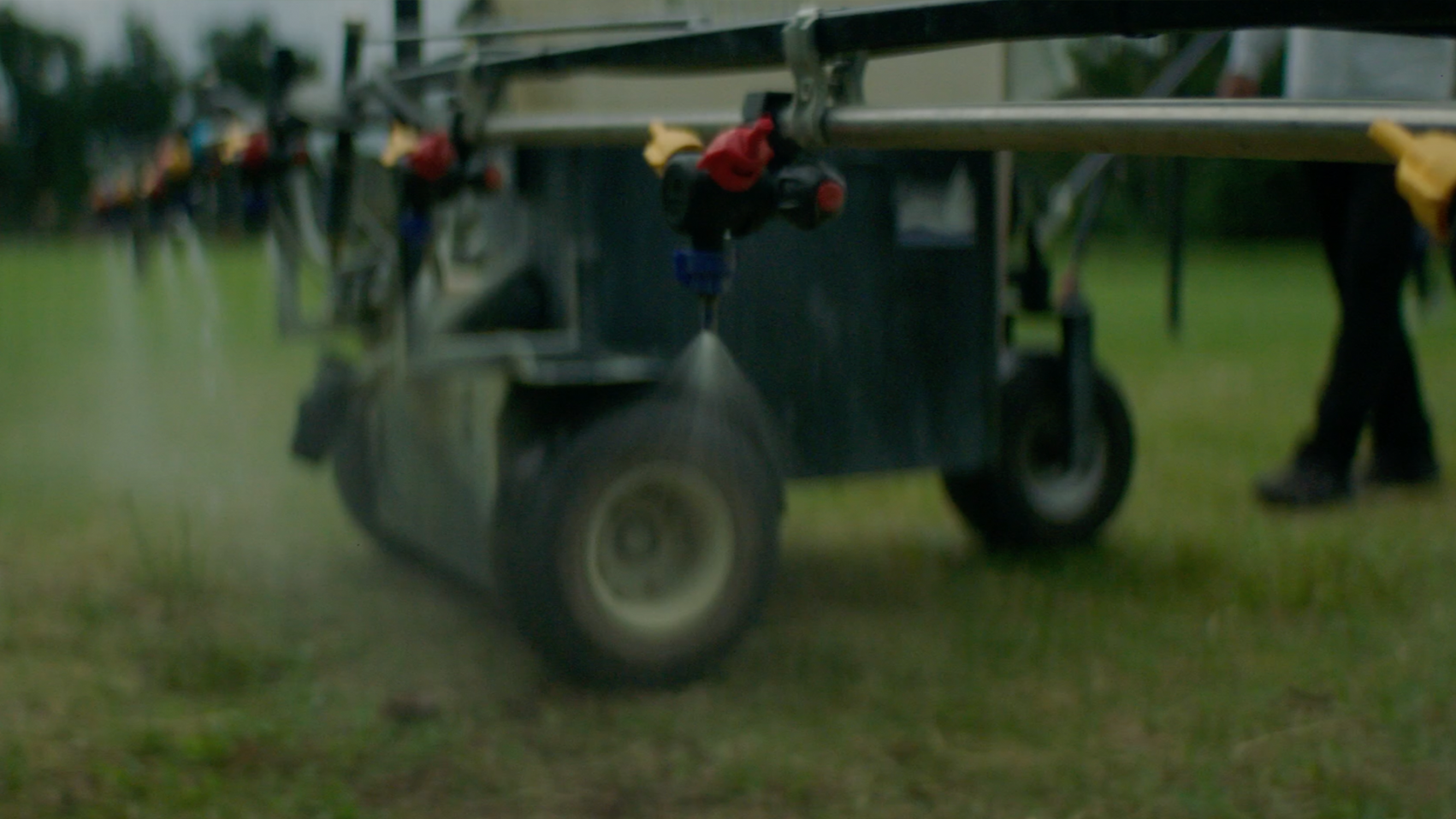 The image shows a close-up of a sprinkler irrigation system on a grassy field, with activity and plants in the background.