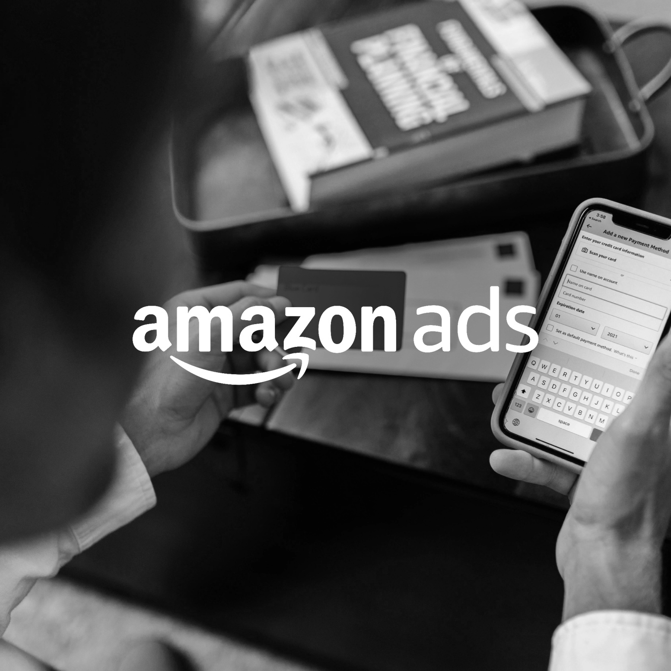 A person viewed from over the shoulder, holding a smartphone and setting up a campaign in the Amazon Ads mobile app. The image is in black and white with the Amazon Ads logo in the center.