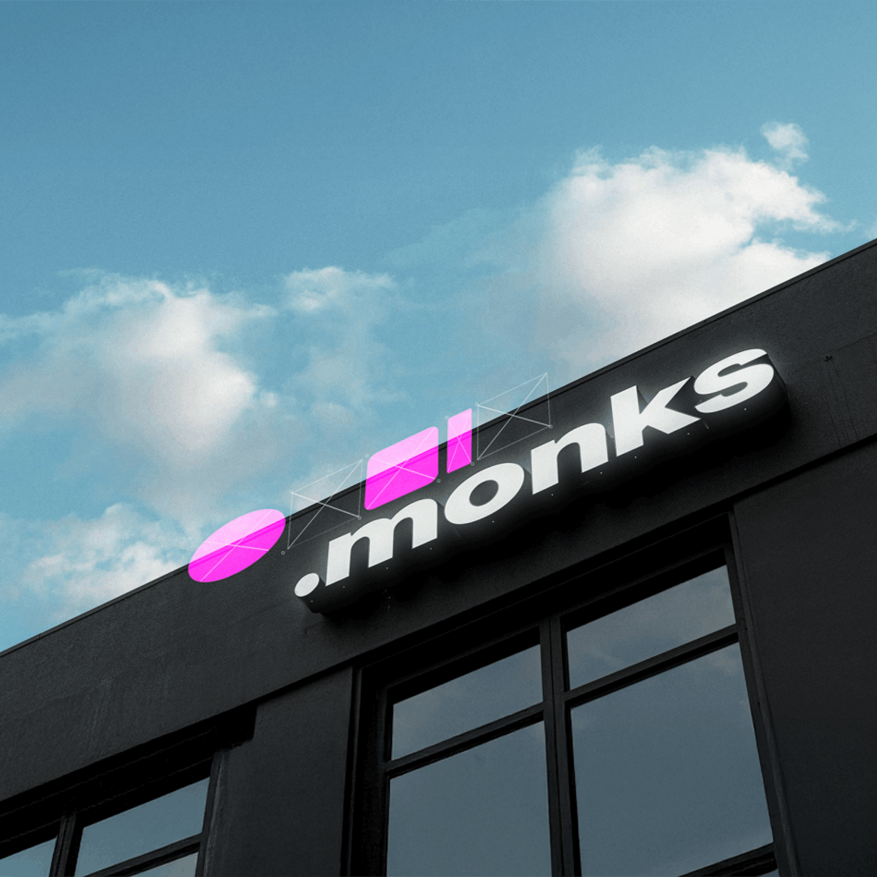 The .monks illuminated logo on the exterior of a modern dark office building against a blue sky.