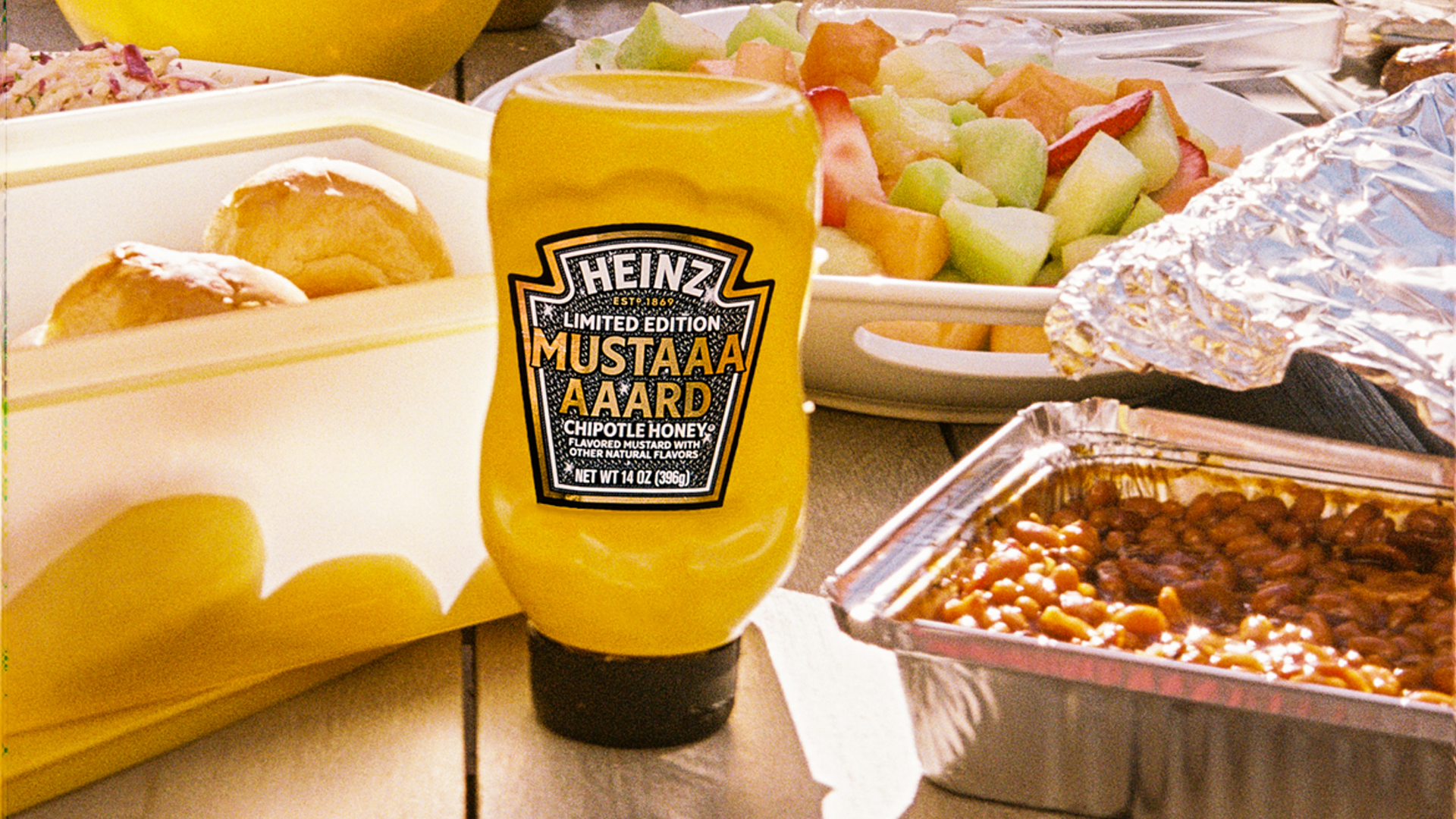 Bright yellow Heinz Limited Edition Mustaaaaard Chipotle Honey bottle, prominently displayed on a wooden picnic table next to a fruit salad, baked beans, and fresh buns, highlighting the perfect summer BBQ condiment.