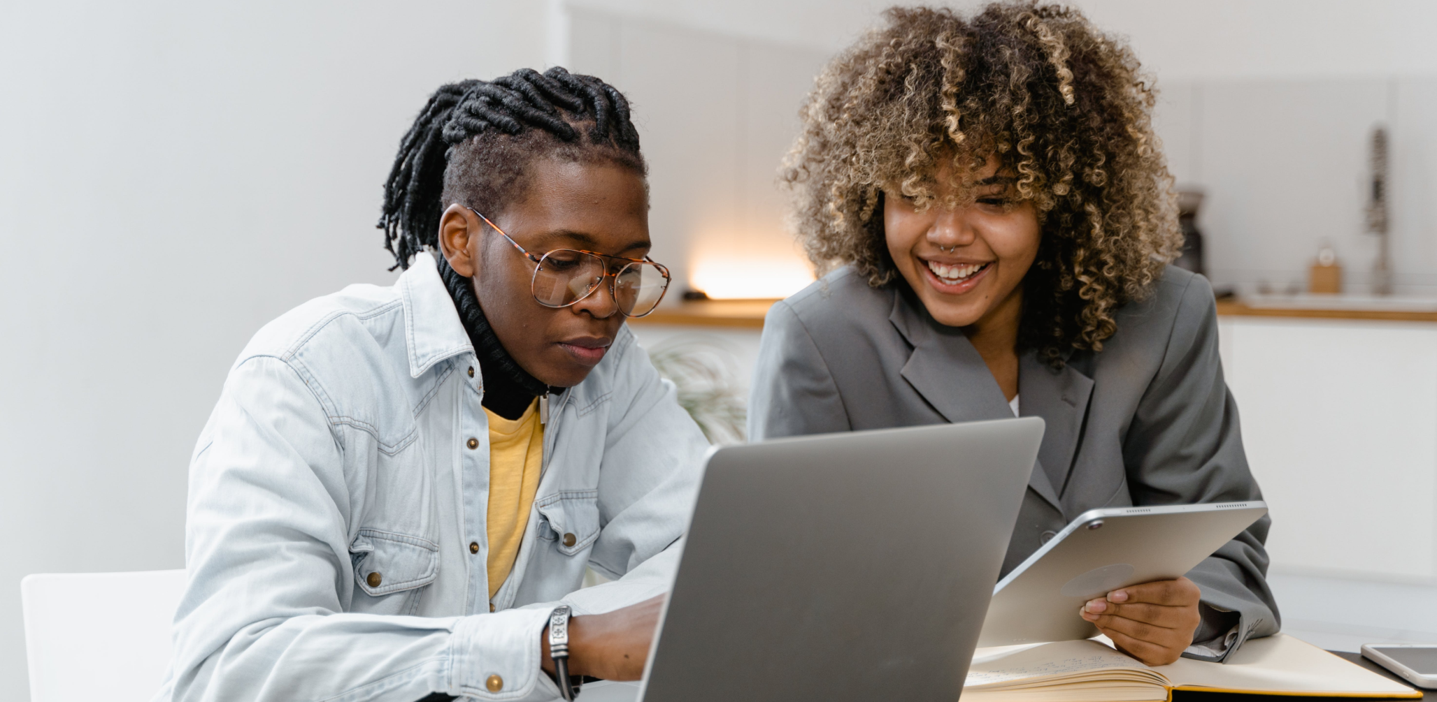 Um homem e uma mulher em um ambiente corporativo olhando para o notebook, ela segura um tablet e está sorrindo 