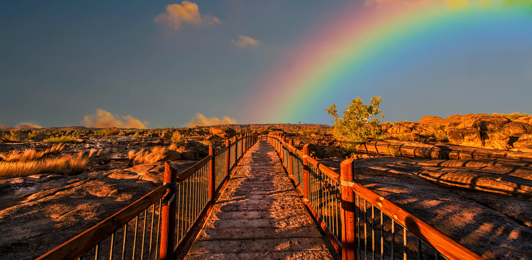 A vibrant rainbow arches over a rustic walkway leading across a rocky landscape bathed in the golden light of sunset. The sky is partly cloudy, enhancing the vividness of the rainbow colors.