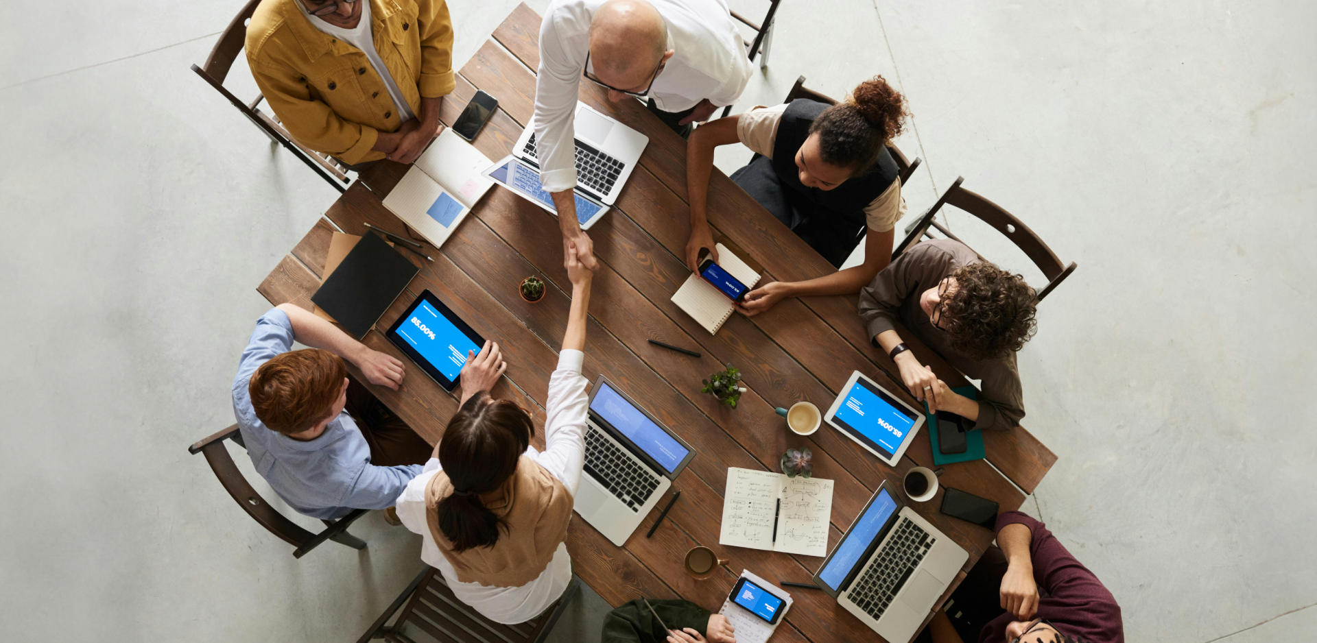 A group of seven people are gathered around a wooden table in a meeting room. They have laptops, tablets, notebooks, and coffee cups in front of them. Two of the individuals are standing and shaking hands over the table, while the others are seated, engaged with their devices or each other. The setting appears collaborative and professional.