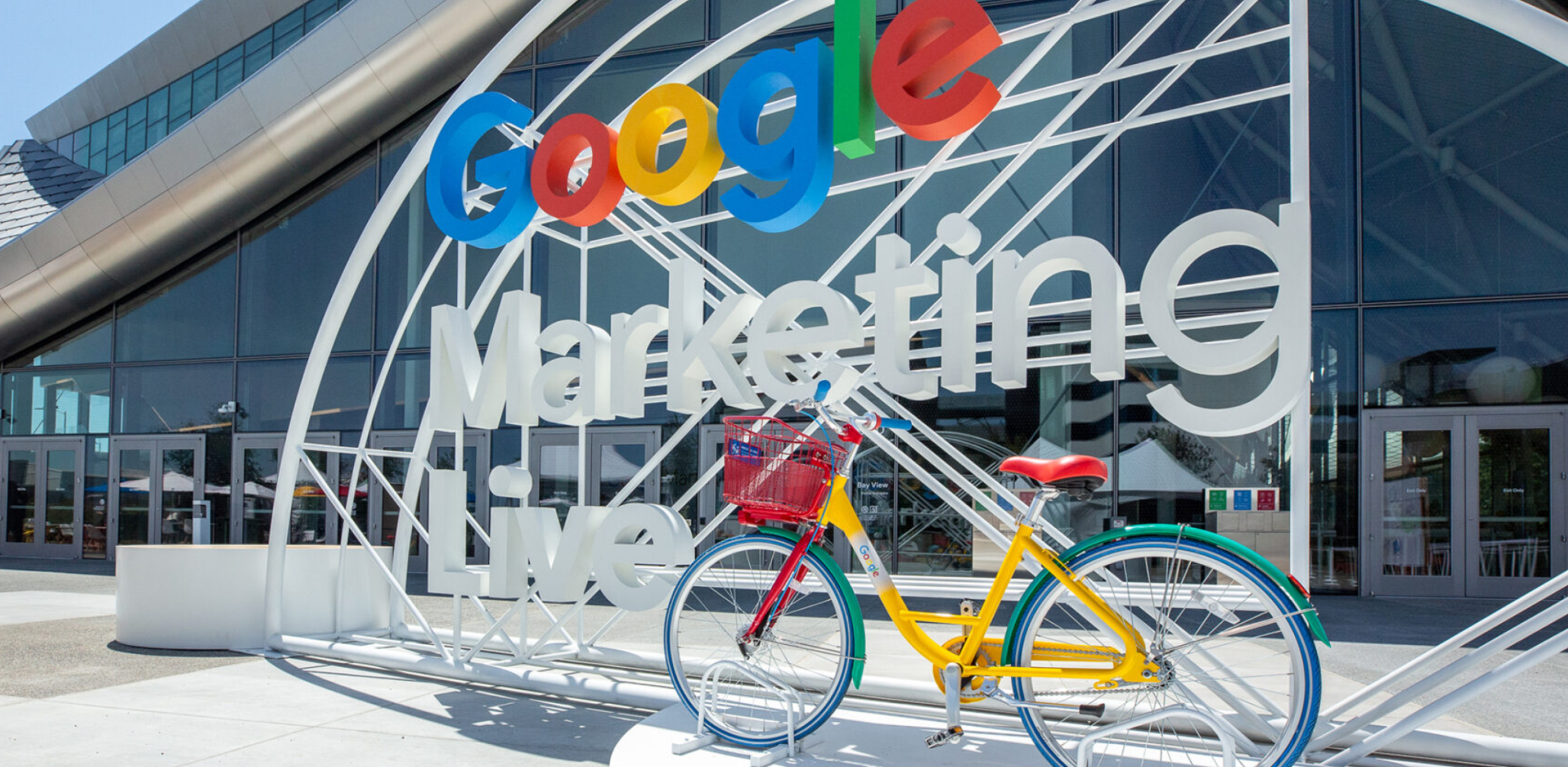 Imagem do evento Google Marketing Live, retratando a entrada principal, destacada por uma grande estrutura com o nome do evento e uma bicicleta colorida característica do Google. O local moderno tem grandes janelas de vidro e arquitetura contemporânea. 