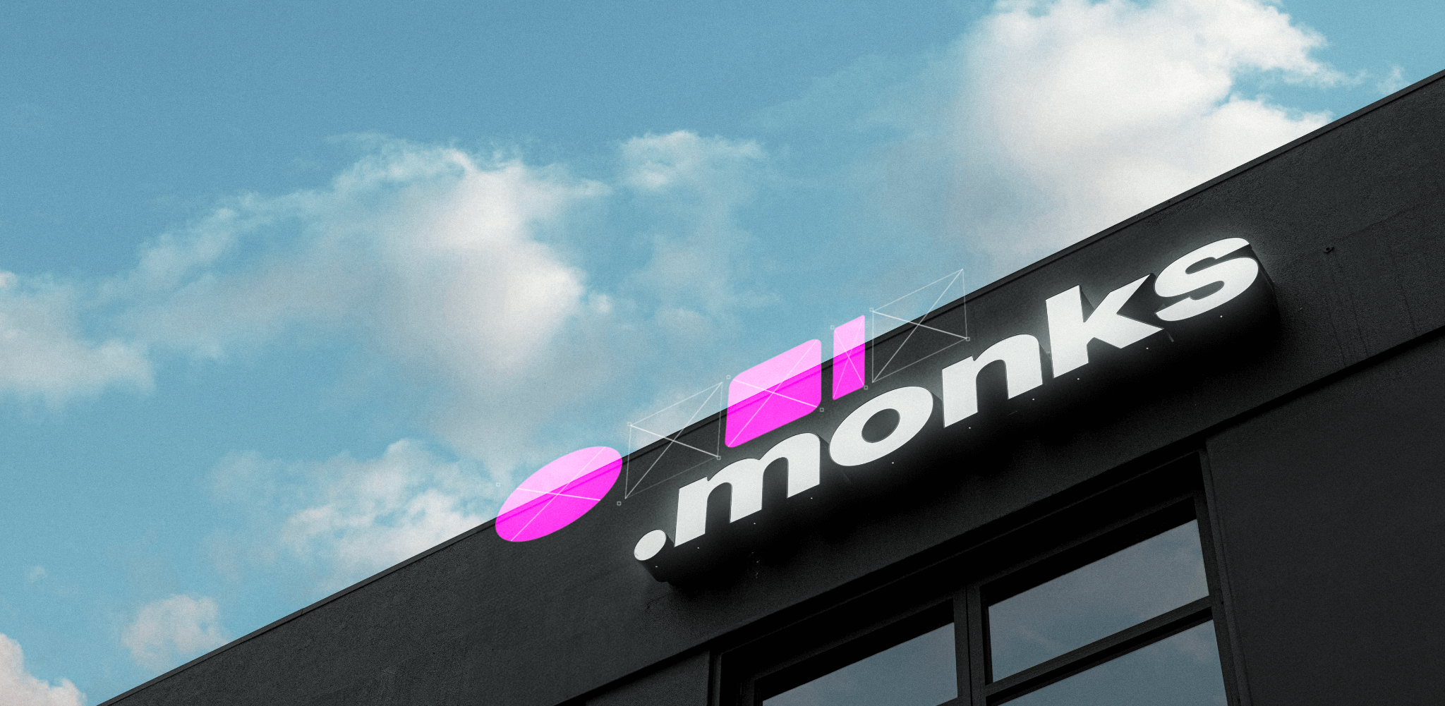 A modern building with a sign that reads ".monks" in white letters on a black background. Above the text are two geometric shapes in pink: a circle and a square. The sky is blue with scattered clouds in the background.