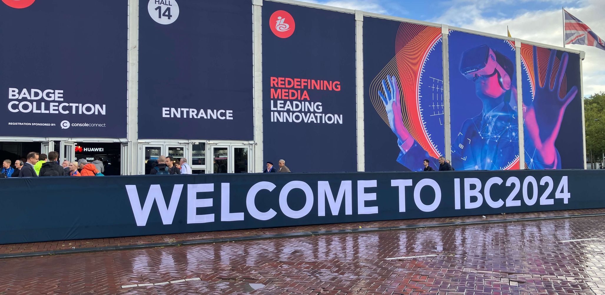 The facade of the IBC convention hall is depicted. Large banners outside display text reading "Badge Collection," "Entrance," and "Redefining Media Leading Innovation." A futuristic image of a person wearing a virtual reality headset is featured. The foreground has a sign that reads "Welcome to IBC2024." People are gathered near the entrance, and a traffic cone is visible on the wet pavement.