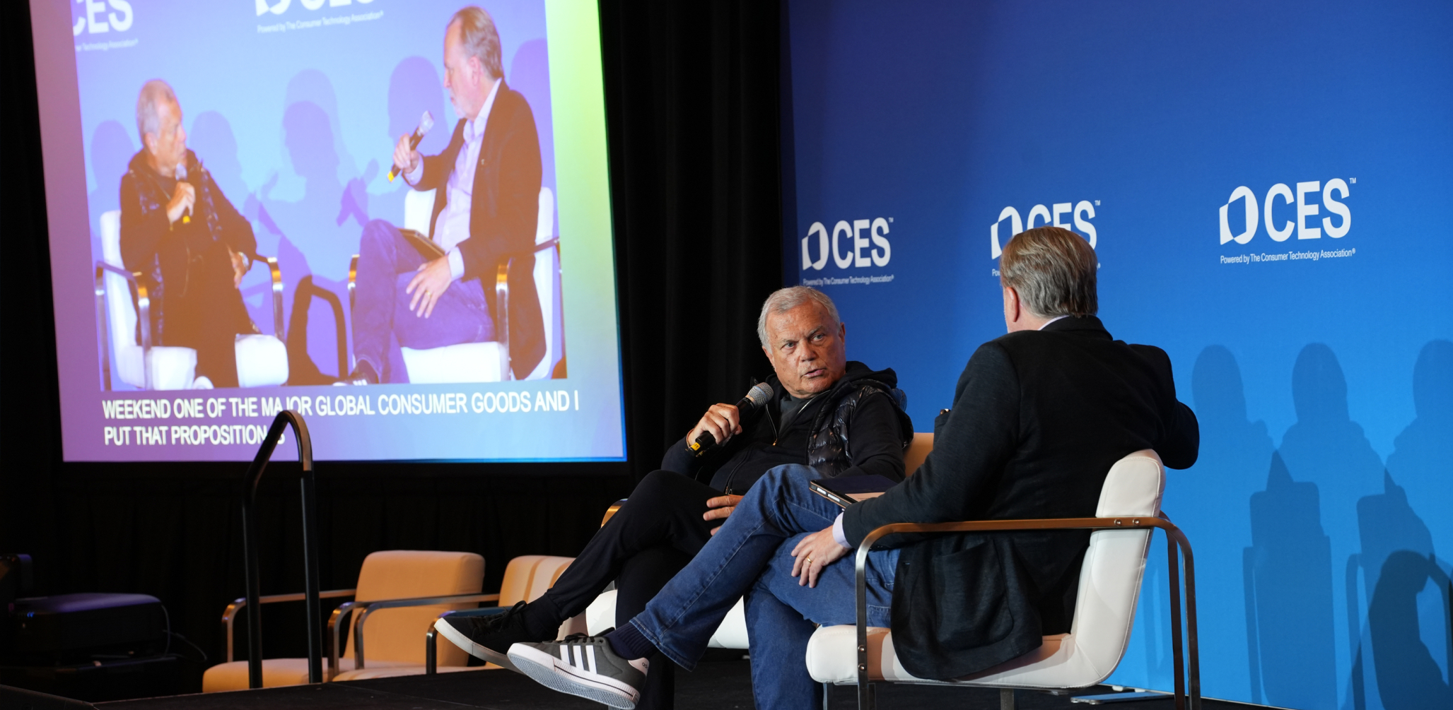 two men sitting on stage at CES with a screen showing them from a different angle in the background