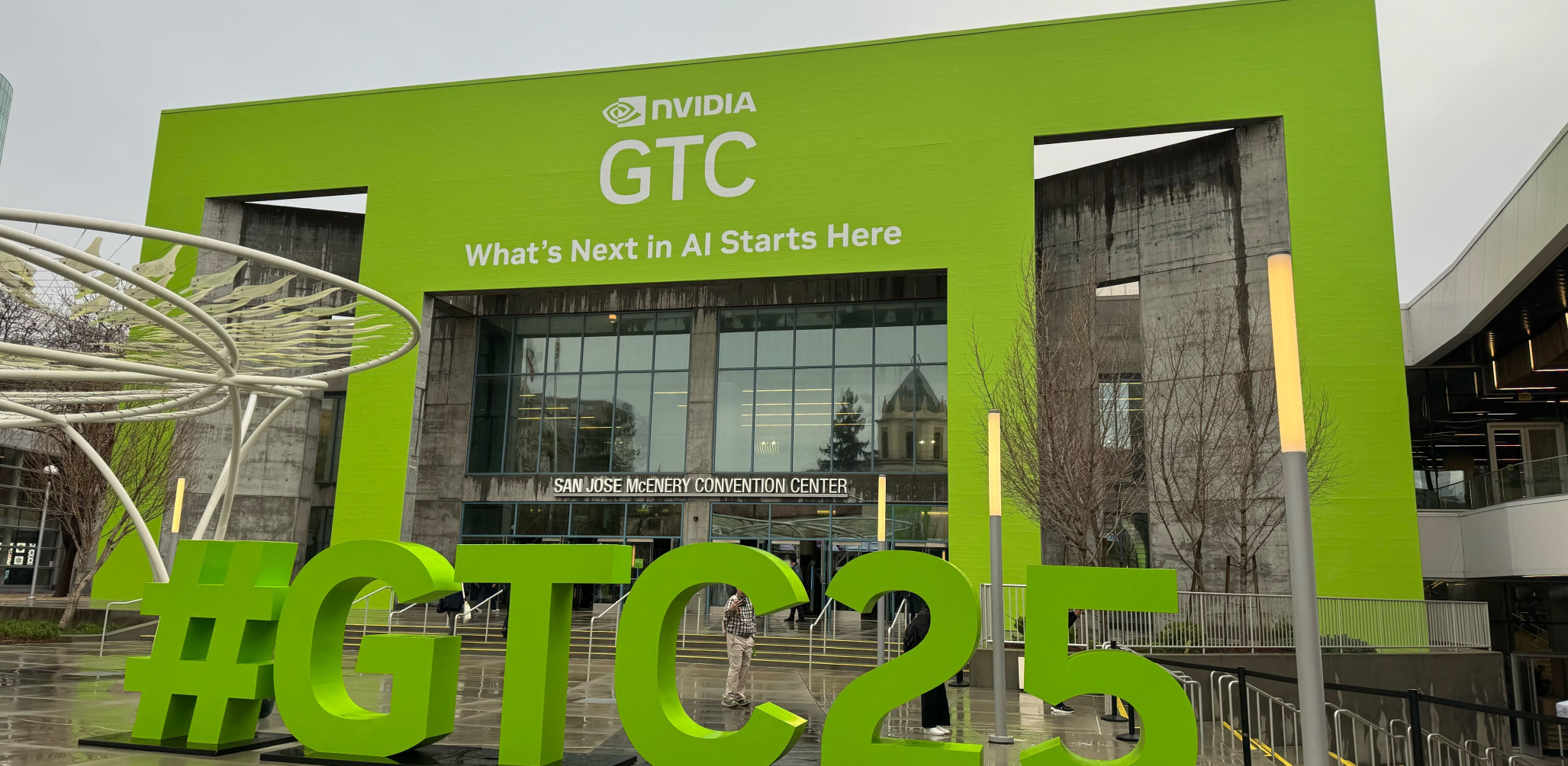 Green building facade that says "NVIDIA GTC: What's next in AI starts here." In the foreground is a sculpture of the hashtag "#GTC25."